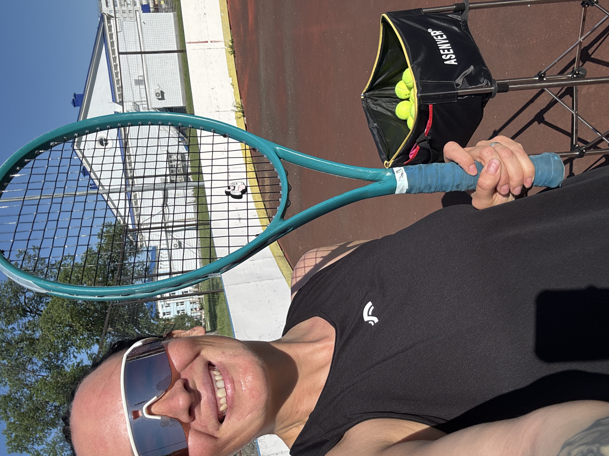 In Chaikovsky, Russia, a woman stands on a tennis court holding a teal tennis racket. She is smiling and wearing sunglasses and a black tank top with a white logo. Behind her, a fence separates the court from buildings and trees. A black basket filled with tennis balls is visible to her right. The court surface is reddish-brown.