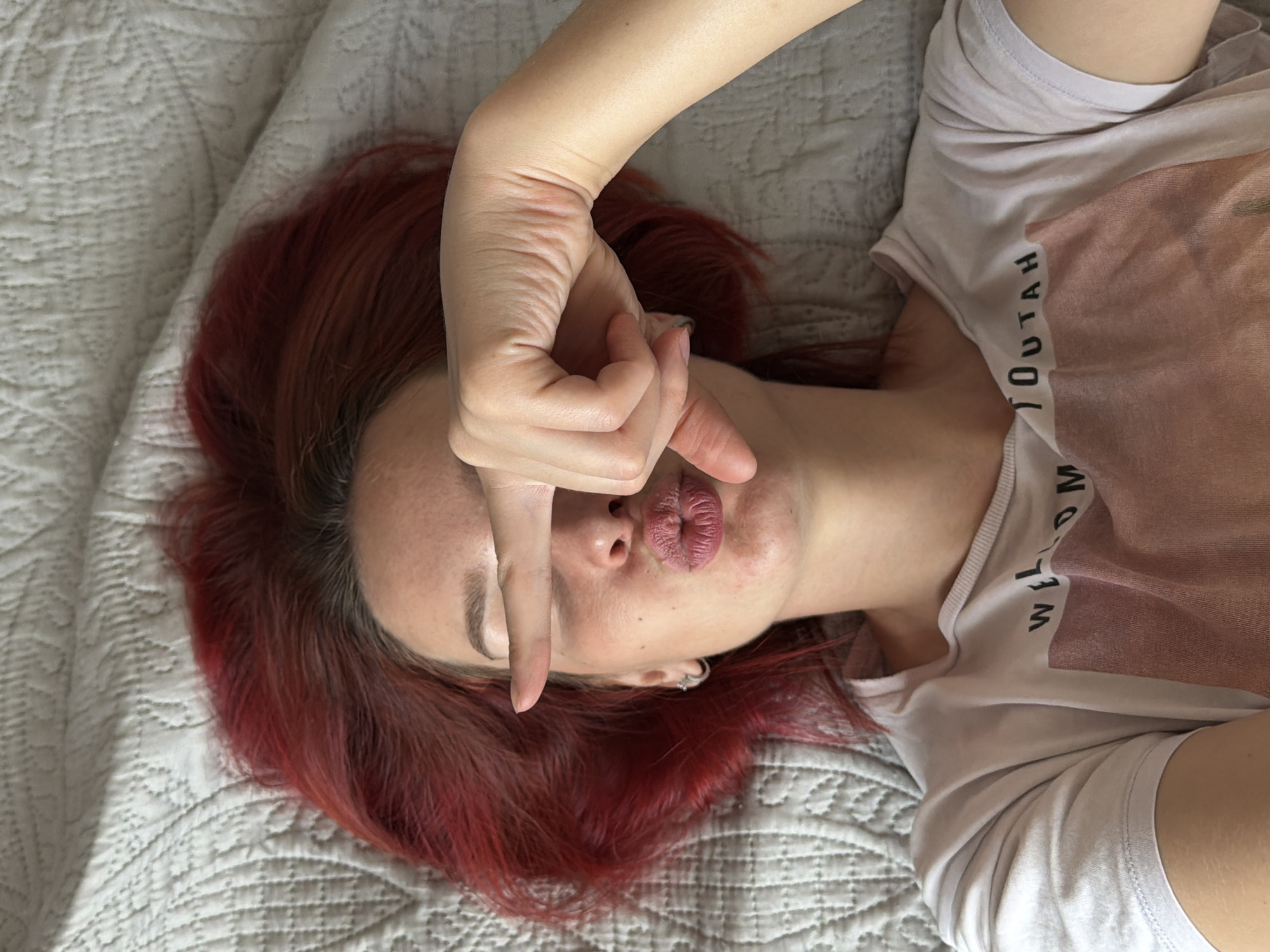 A woman lies on a gray quilted bedspread. Her hair is red. She wears a white T-shirt with the word "YOUTAH" written vertically down the right side. Her right hand is held up, with the index and middle fingers extended vertically in front of her closed left eye. Her lips are pursed. The location is unspecified.