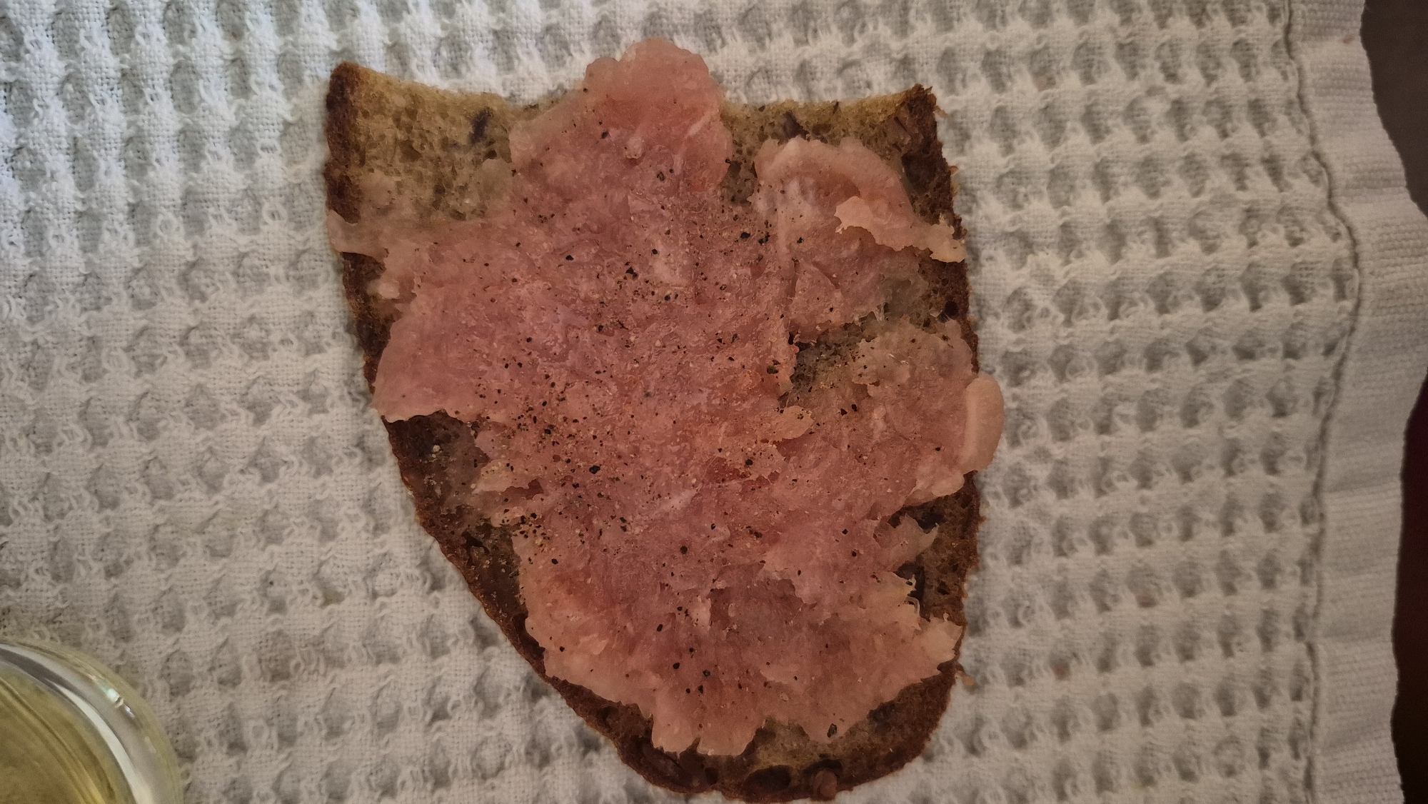 A close-up, overhead shot shows a slice of dark bread topped with a pinkish spread and sprinkled with black pepper. The bread is irregularly shaped and appears to be a single slice from a larger loaf. The spread has a coarse, shredded texture. The bread and topping are placed on a white waffle-weave textured cloth. In the upper left corner, a portion of a clear glass containing a pale yellow liquid is visible. The scene is lit from above and appears to be indoors. The image does not contain any information that would allow it to be located in Krauja, Latvia.