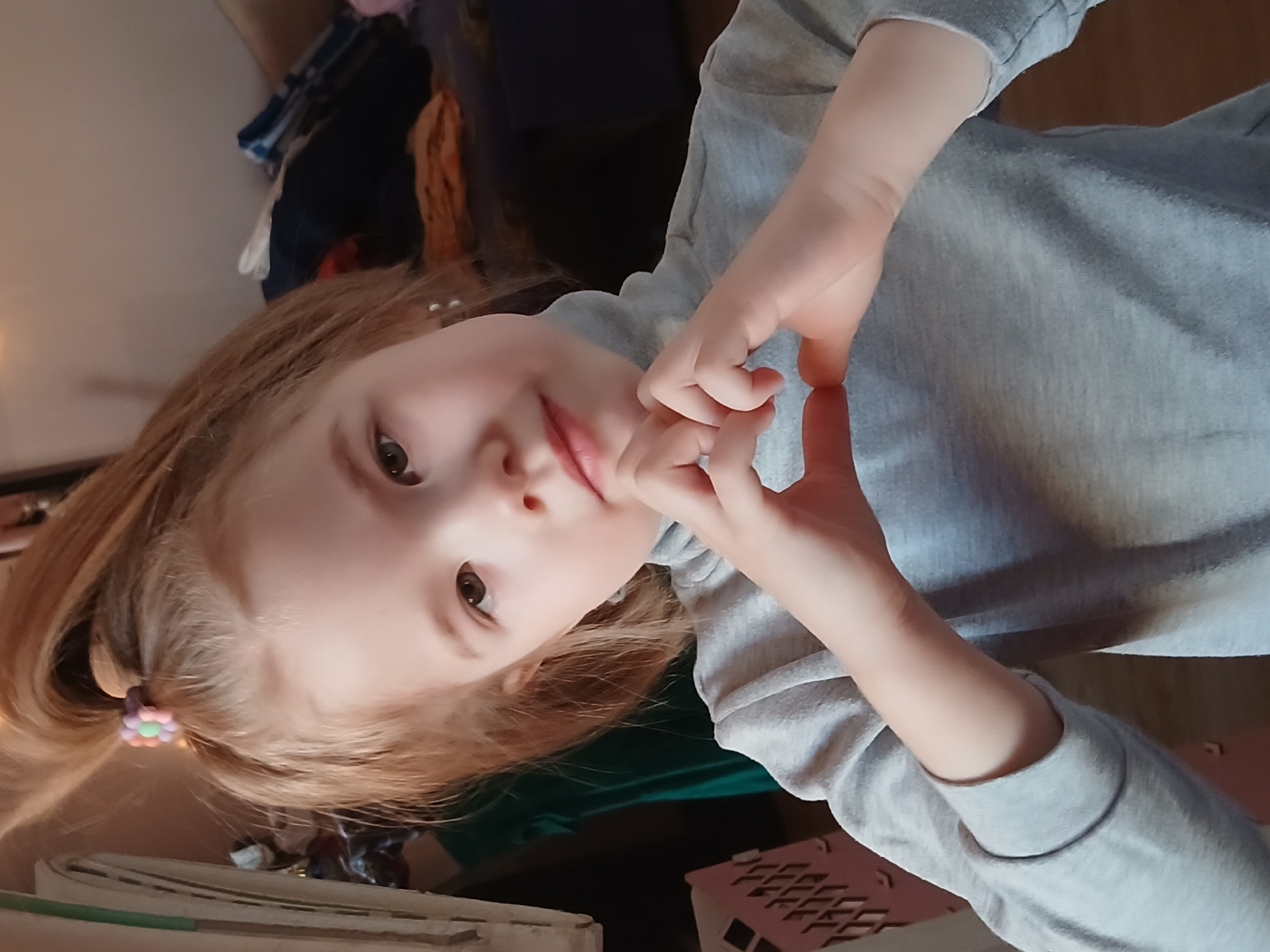 A young girl with fair skin and light brown hair is making a heart shape with her hands. She has a light grey long-sleeved shirt on. The background includes a dark blue object and another object with a grid pattern. The location is in the city of Chaikovsky, Russia.
