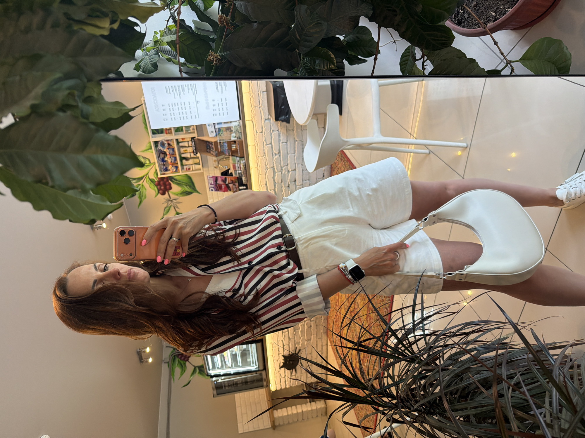 The image shows a woman taking a selfie in a cafe. She is wearing a striped shirt, white shorts, and white sneakers, and she is carrying a white handbag. The cafe appears to be modern and well-lit, with white brick walls, a coffee bar, and seating areas. There are plants in the foreground and background, adding a touch of greenery to the scene. The woman is looking at her phone, which is held up to take the picture. The overall impression is of a casual and relaxed atmosphere. However, there is no information in the image that suggests the location is in Birobidzhan, Russia.