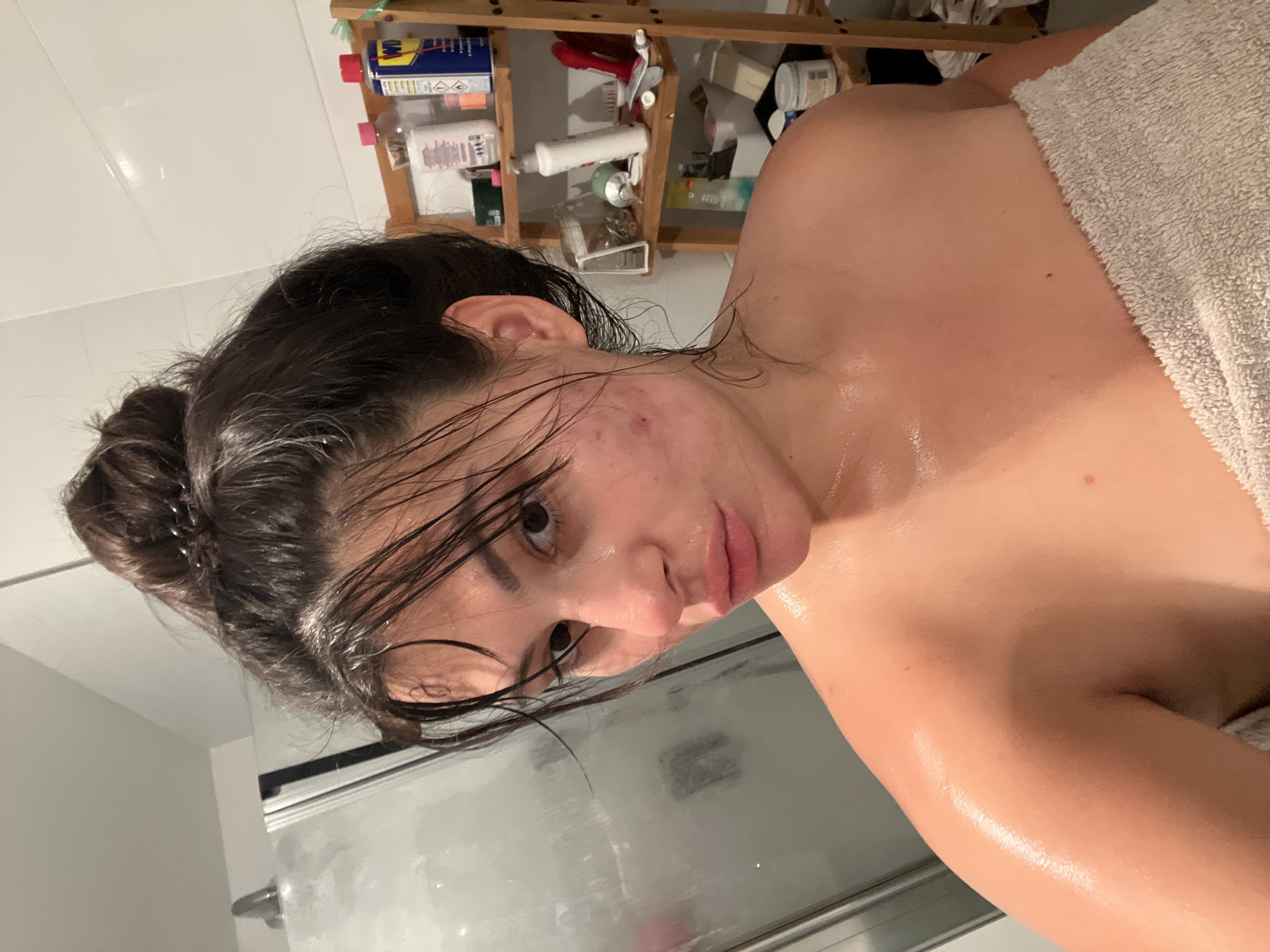 In a bathroom, a young woman with her hair in a bun and wrapped in a towel, poses for a selfie. Her face is glistening with water and she has several acne blemishes on her cheeks. To the left of the woman is a shower stall with a glass door. To the right of the woman, a wooden shelf unit is visible, filled with various toiletries and cleaning supplies. The image was taken in Kiryat Bialik, Israel.