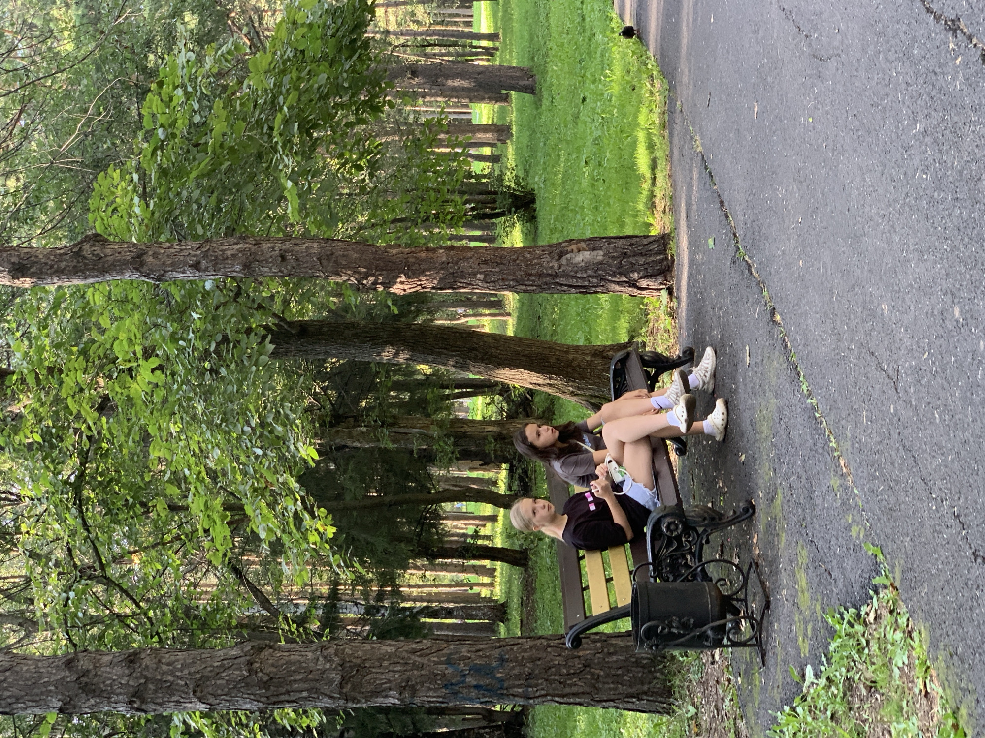 Two young females are seated on a park bench in a wooded area. The female on the left has blonde hair pulled back and is wearing a black t-shirt and white shorts. The female on the right has dark brown hair and is wearing a grey t-shirt and white shorts. Both are wearing white sneakers and white socks. The bench is made of dark metal with a yellow wooden seat. A black trash receptacle with ornate metalwork is to the left of the bench. The ground in front of the bench is a paved path, and to the right of the path is a well-maintained grassy area. Behind the bench, a dense forest of tall trees with green foliage creates a dappled sunlight effect on the grass. The location is identified as the City Park in Prokopyevsk, Russia, based on the provided context. A small black bird is visible on the path to the right of the bench.