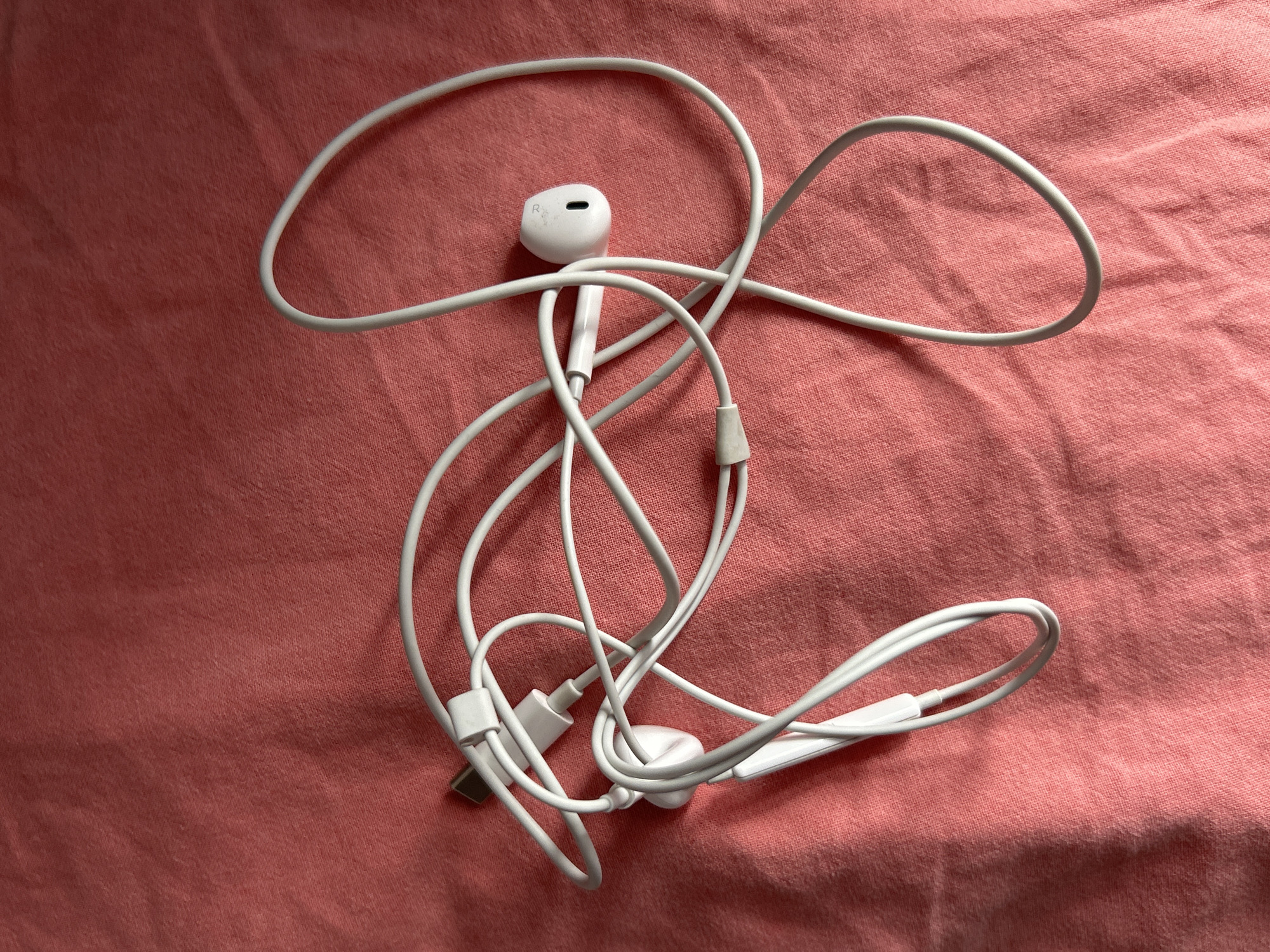 A white wired earphone set with a Lightning connector is sprawled across a pink, textured fabric surface. The earphone set is tangled, with the cord forming various loops and curves. One earbud, marked with an "R" for right, is clearly visible. The fabric appears to be a bedsheet or similar material. There are no people or any indications of a specific location in Moscow, Russia.
