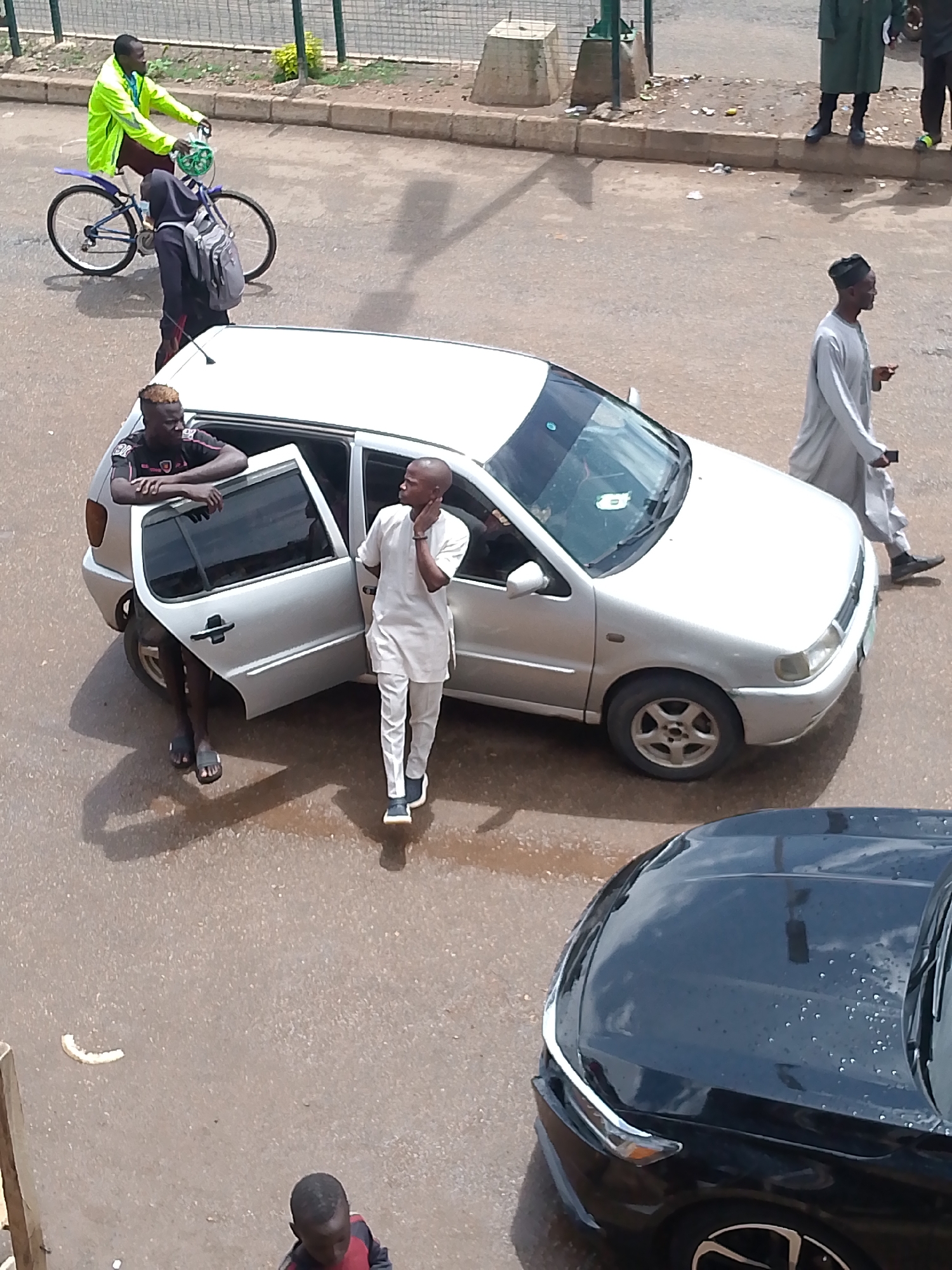 In Bukuru, Nigeria, a man in a bright yellow jacket rides a bicycle, with a person in a dark hooded garment and a backpack walking beside him. A silver car is parked, its front passenger door open, with one man leaning on the car and another standing near the open door. Another man in light-colored clothing walks past the silver car. In the foreground, the hood of a black car is visible. The scene takes place on an asphalt road, with a sidewalk and a metal fence in the background.