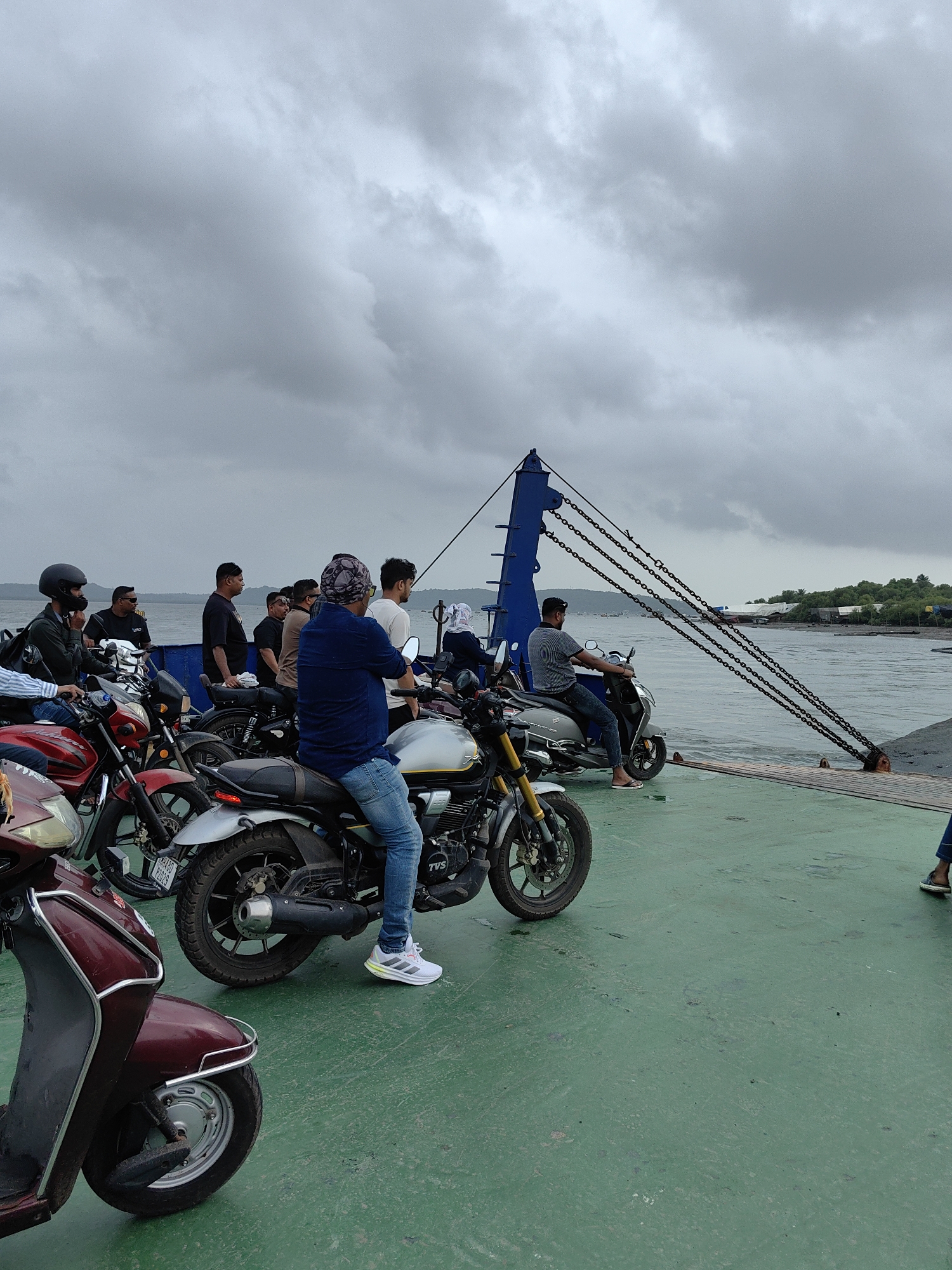 A group of people, mostly men, are gathered on a ferry with their motorcycles and a scooter. The ferry is painted green on its deck. A large blue structure with thick chains is visible on the right side of the ferry, likely part of its loading ramp or mechanism. The sky is overcast with grey clouds. In the background, a body of water with some boats is visible, and on the far shore, a treeline and buildings suggest a coastal or riverside location. The people are standing or sitting on their vehicles, some are looking around, while others appear to be conversing. One person in the foreground is wearing a blue shirt and jeans, sitting on a silver motorcycle. Another person in the middle ground is wearing a grey t-shirt and jeans, sitting on a scooter. A red scooter is parked in the lower left corner of the frame. The scene depicts a mode of transport, likely a ferry service for vehicles and passengers. The location is identified as Mira-Bhayander, India.