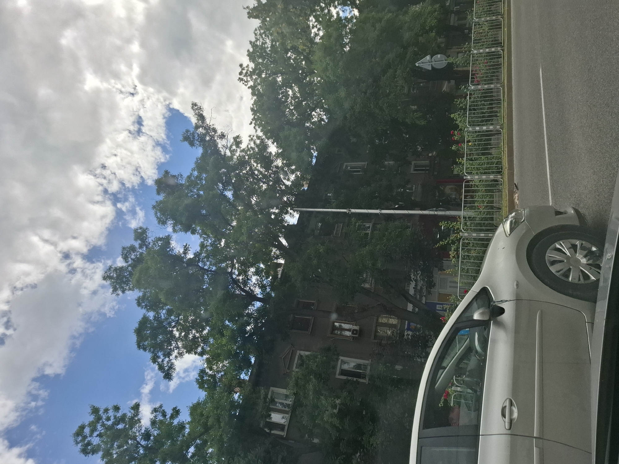 The image depicts a street scene in a city, likely during the daytime under a partly cloudy sky. A silver car is visible in the foreground, partially obstructing the view of the surrounding environment. Behind the car, a building with multiple windows and balconies is partially obscured by dense green trees. A metal fence lines the sidewalk in front of the building, with some red flowers visible behind it. A street sign is also present on the right side of the image. The overall impression is of an urban residential area with significant greenery.