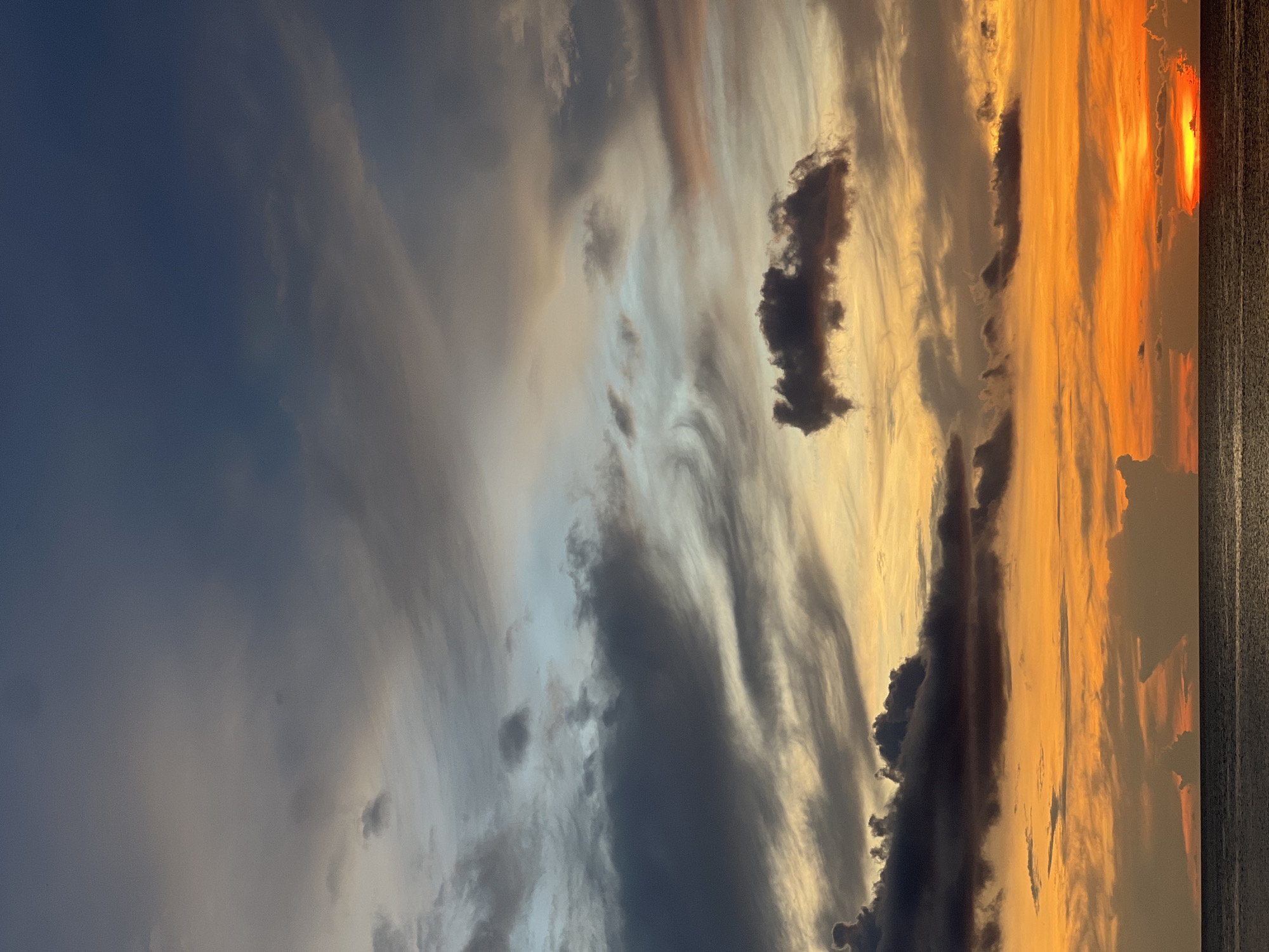 A sunset fills the sky with a gradient of colors from dark blue on the left to oranges and yellows on the right. Clouds of varying sizes and shapes are scattered throughout, with darker, more defined clouds closer to the horizon. The sun is visible on the far right, near a dark vertical strip that could be the edge of a building or landscape. The image appears to be taken in an urban environment at twilight. The location is Unknown, Unknown.