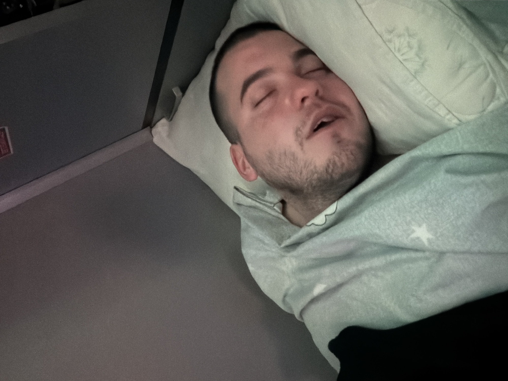 A man is asleep on a bed, presumably on an airplane. He is lying on a pillow and covered with a blanket with star patterns. The setting appears to be inside an aircraft cabin, based on the wall paneling in the background. There is no contextual evidence indicating the specific location is Varna, Bulgaria.