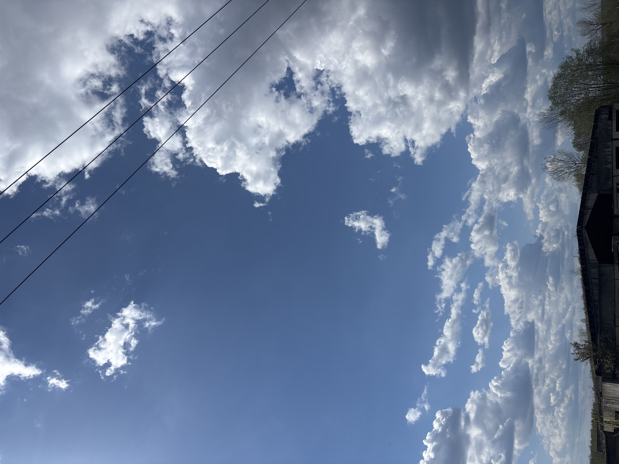 The image shows a partly cloudy blue sky in Раздолье, Russia. Utility wires are visible in the upper left corner of the frame. The right side of the image includes the edge of a building and trees. The overall scene depicts a bright, daytime sky with clouds.