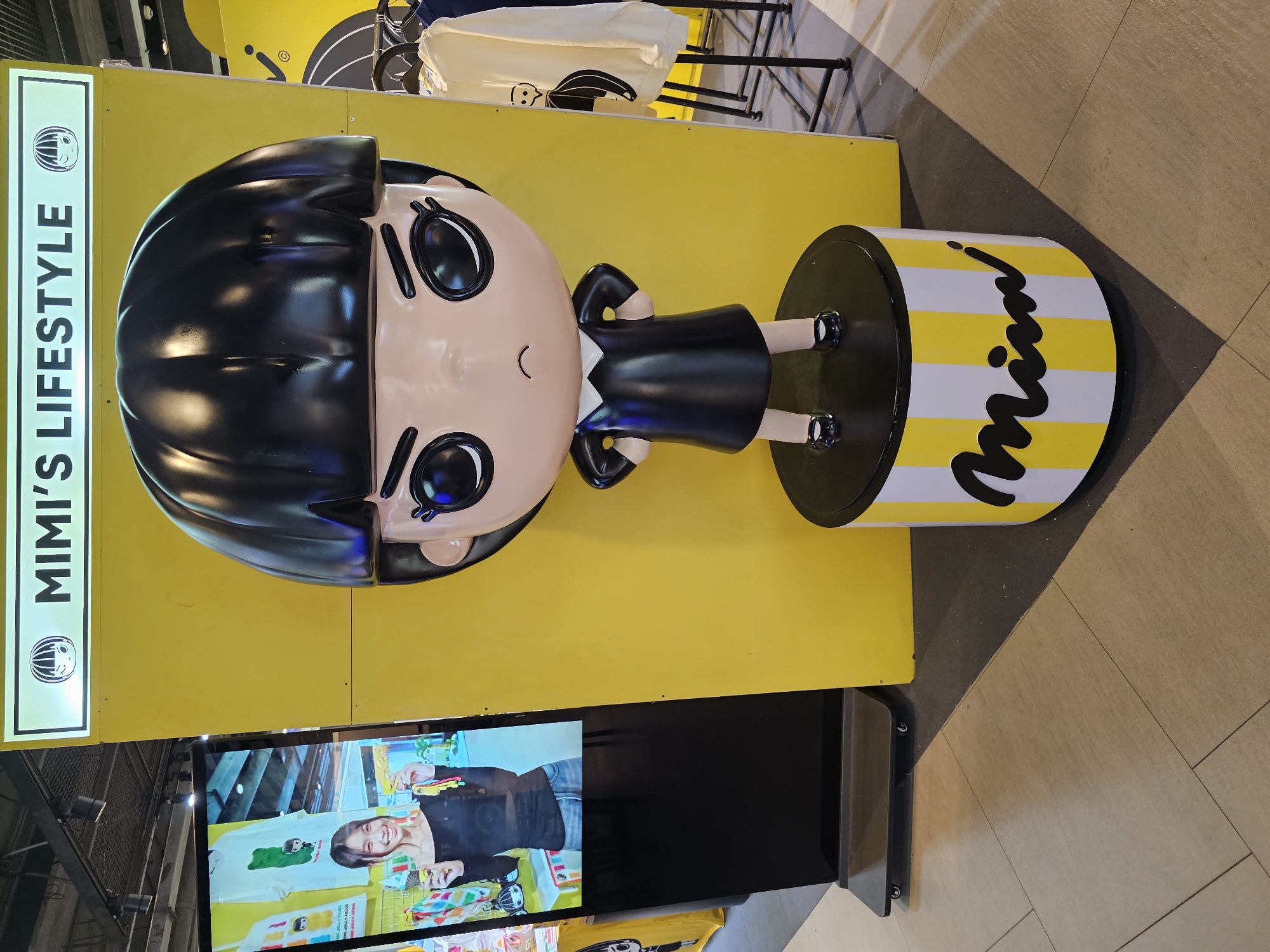This is a full shot of a large, stylized doll figure in a black dress, standing on a cylindrical display base with yellow and white stripes and the word "Mimi" in black script, located in an indoor retail environment. The doll has a large head with a black bob haircut, large black eyes with thick eyebrows, and a small, frowning mouth. Its arms are akimbo. The background consists of a large, bright yellow wall with a sign at the top that reads "MIMI'S LIFESTYLE" with a cartoon illustration of a girl's face. To the left of the doll is a tall, black digital display screen showing a woman holding an ice cream cone and wearing a black off-the-shoulder top and jeans. Further to the right, clothing items hang on racks. The flooring is tiled with a wood-like pattern. This scene is located in the city of Bangkok, Thailand.