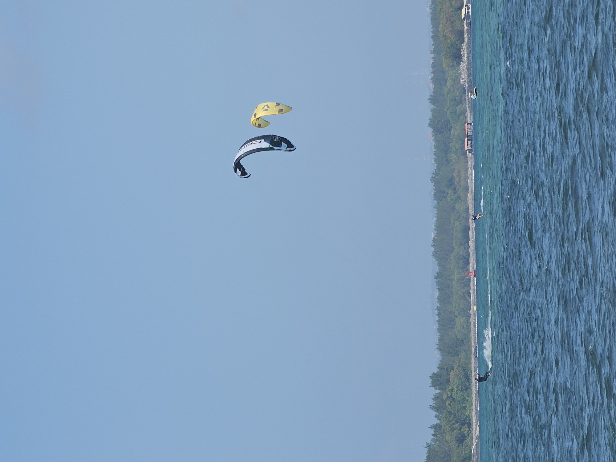 The image depicts a bright, sunny day with a clear blue sky above a body of water. Two kitesurfers are visible in the distance, one with a black and white kite and the other with a yellow kite. One kitesurfer is closer to the foreground and is actively riding on the water, creating a spray behind them. The water is a deep blue with visible ripples. Along the horizon, there is a lush green treeline, indicating land with dense vegetation. Several small structures, possibly buildings or huts, can be seen nestled within the trees. Further out on the water, there are a few small boats or buoys. This scene is likely located in or near Denpasar, Indonesia, a city known for its coastal areas and water sports. The overall impression is one of an active and scenic coastal environment on a pleasant day.