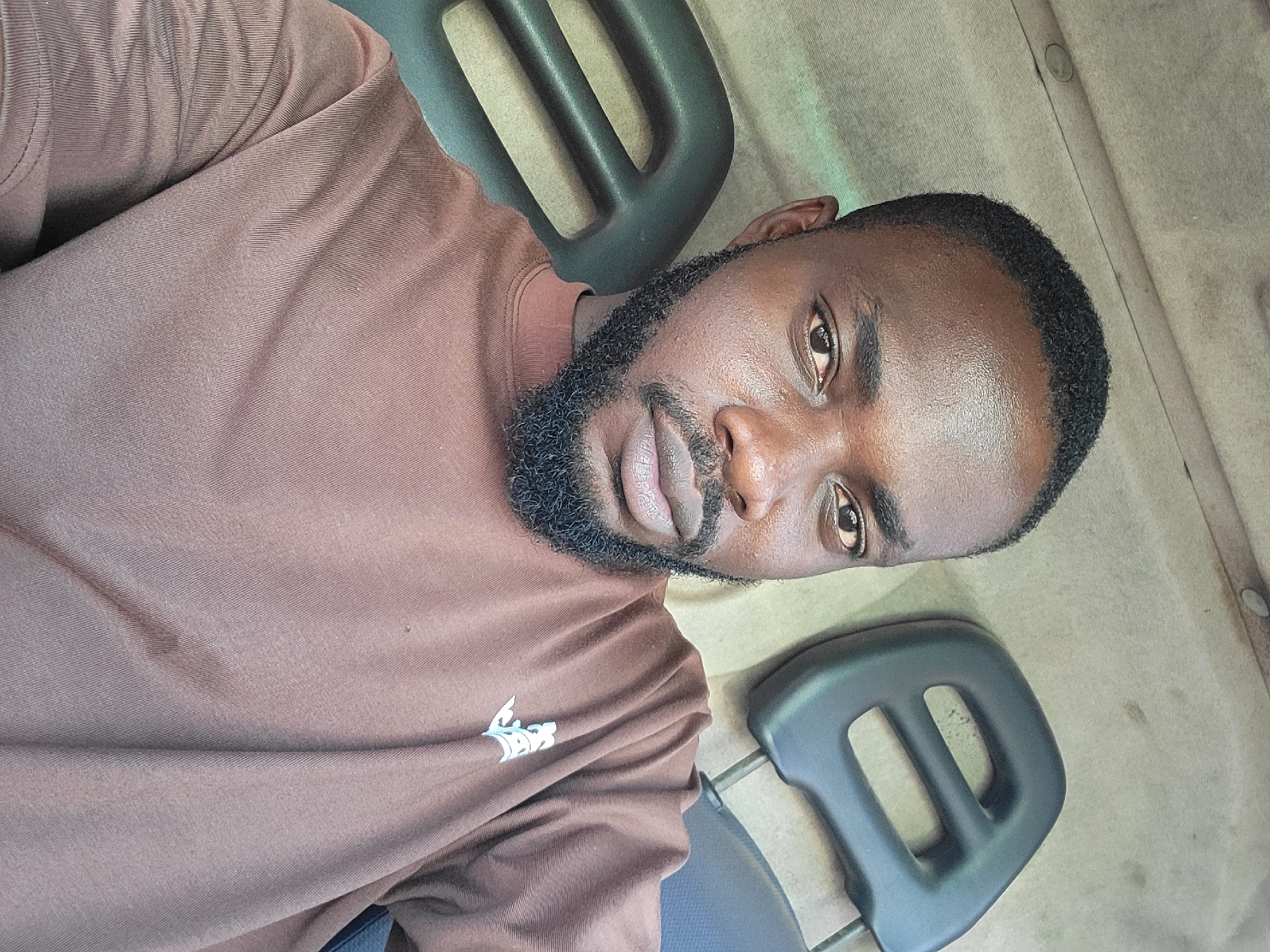 A medium, angled shot of a Black male with a trimmed beard and short dark hair, wearing a brown long-sleeved shirt with a small, white logo on the front left. He's seated in what appears to be a vehicle cabin, judging by the two dark headrests on metal posts positioned in the background. The interior is a tan color with minor blemishes. The location in Jos, Nigeria cannot be confirmed without further visual or contextual clues.
