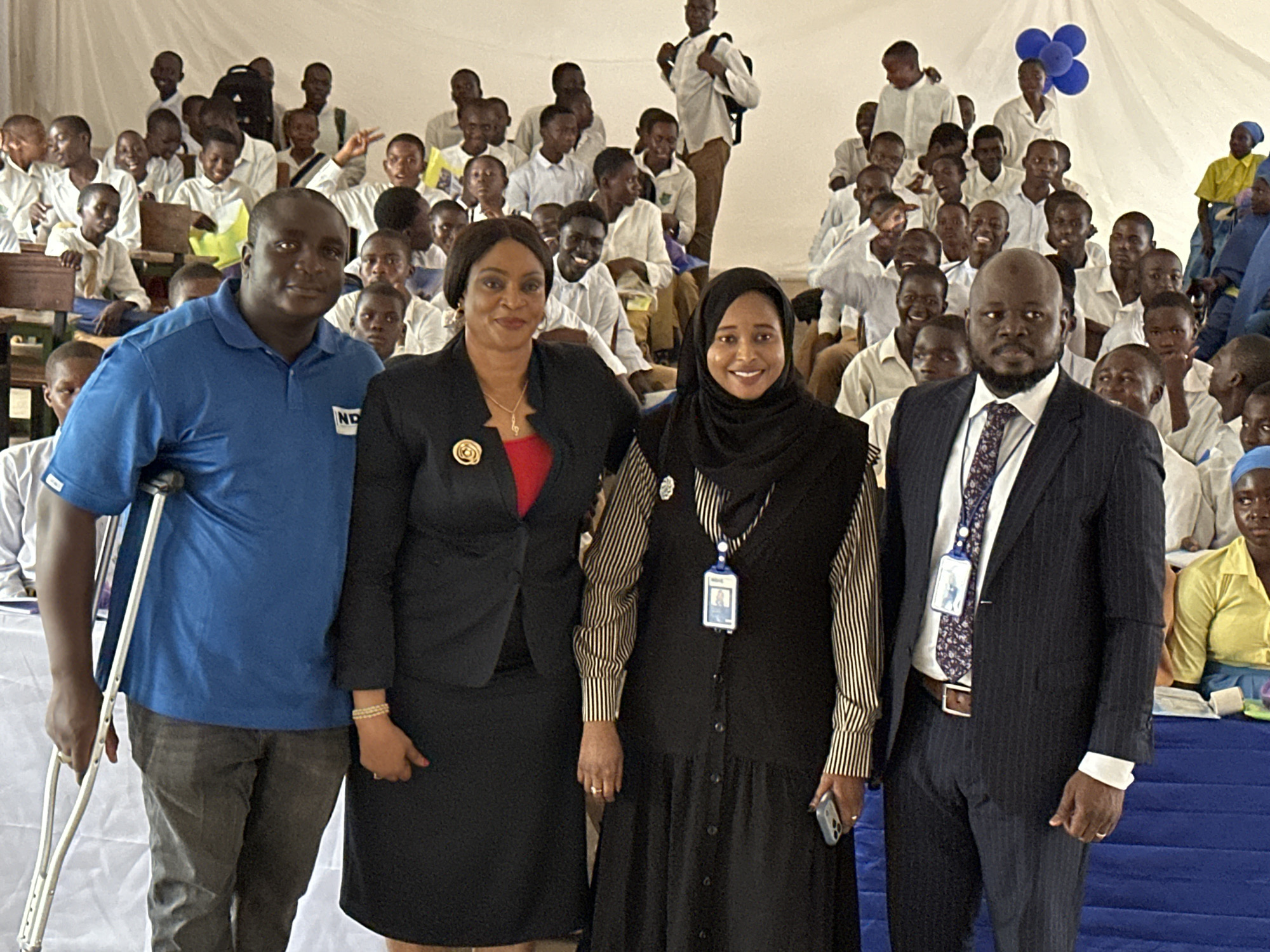 The image captures a group of four adults posing in the foreground, with numerous young people, likely students, seated in rows behind them. The setting appears to be an indoor event, possibly a school assembly or presentation, in Unguwan Dosa, Nigeria. White fabric drapes form the background and ceiling, suggesting a tented or specially decorated space.
From left to right, the adults include a man wearing a blue polo shirt with "ND" visible on it and grey trousers, holding a crutch under his left arm. Next to him is a woman in a black blazer over a red top and a black skirt, adorned with a gold brooch. The third adult is a woman wearing a black hijab, a striped shirt under a black button-down dress or jumper, and an ID badge around her neck. The fourth person is a man in a dark pinstripe suit, white shirt, and patterned tie, also wearing an ID badge. All four adults are smiling at the camera, conveying a positive mood.
In the background, a large group of young people, predominantly boys, are seated on wooden desks and benches. Most are wearing white long-sleeved shirts, appearing to be school uniforms. Many students are also smiling and engaged, looking towards the front. Blue balloons are visible in the upper right corner, and a blue-draped table is seen in the bottom right foreground. The lighting suggests it is daytime. One of the ID badges has an unreadable photo and text.