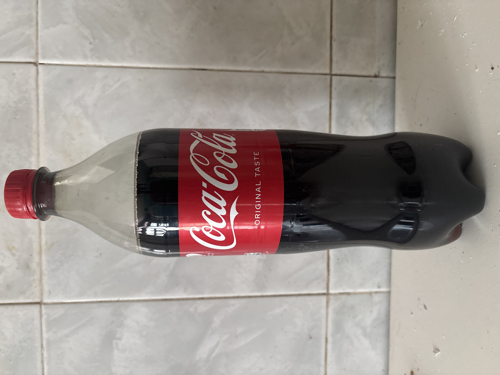A clear plastic bottle of Coca-Cola is positioned vertically in the center of the frame. The bottle is filled with a dark brown liquid, and a red cap is visible at the top. The Coca-Cola logo, in white script, is prominently displayed on a red label around the bottle's body. The background consists of a tiled wall, with off-white rectangular tiles. The bottle rests on a white surface that appears to be a countertop, with some small debris scattered on it. The lighting suggests an indoor environment. This scene is not located in the city of Campomarino, Italy.