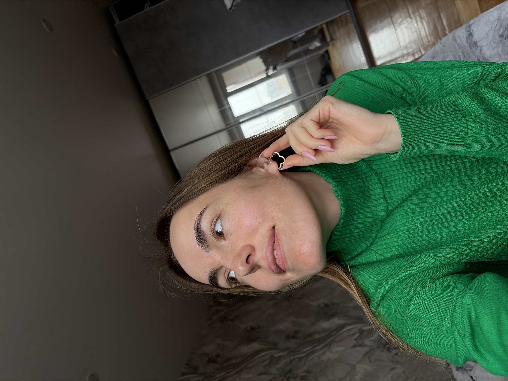 A woman with light brown hair and a green sweater is holding a black clover-shaped earring near her left ear. The background shows a room interior with a gray wall, mirrored cabinet, and parquet floor. It is not possible to determine if the location is Izhevsk, Russia, based on this image alone.