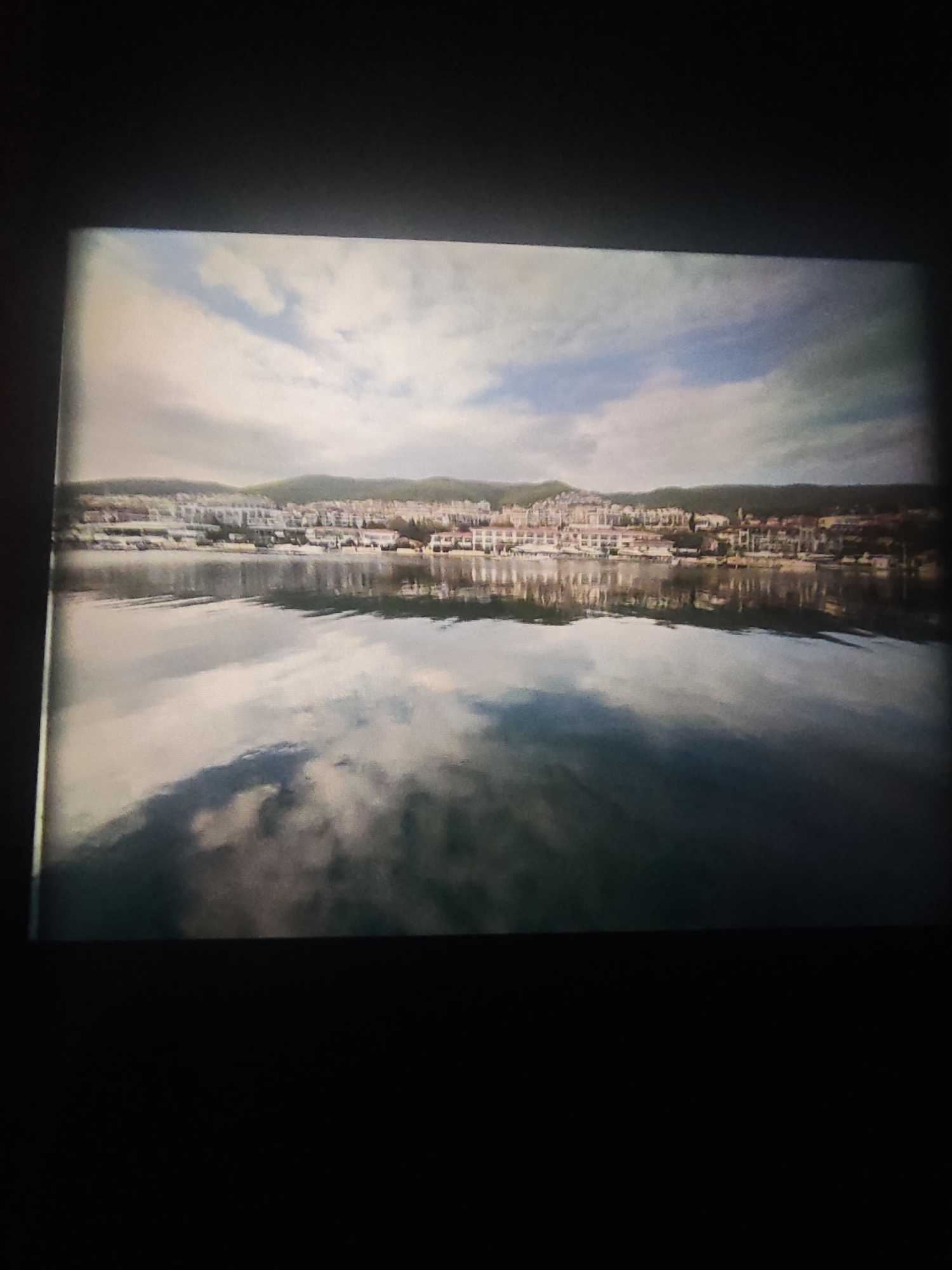 The image shows a body of water with the reflection of clouds in the water. On the opposite shore are buildings, possibly houses, and a hillside behind them. The sky is cloudy. The location cannot be determined without additional information.