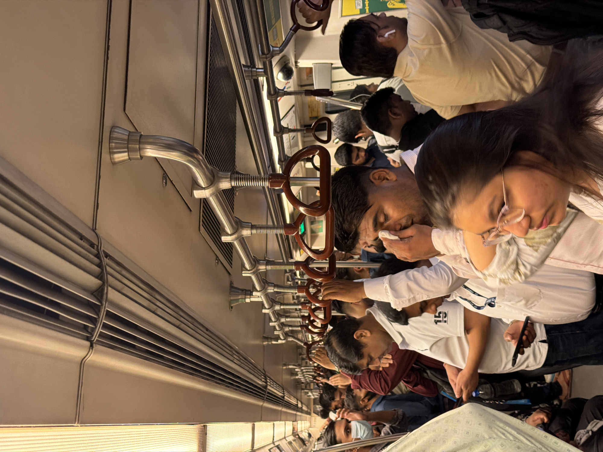 The image depicts the interior of a crowded Delhi Metro train carriage. Numerous passengers, primarily adults, are standing in close proximity, with many holding onto overhead handrails for support. The lighting suggests it is either daytime or a well-lit underground environment. Several individuals are looking at their mobile phones, while others are engaged in conversation or simply enduring the crowded conditions. One man in the foreground is seen wiping his face with a tissue. The environment is utilitarian, consistent with public transportation, featuring metal poles, hand grips, and ventilation systems along the ceiling. The density of people indicates peak travel hours.