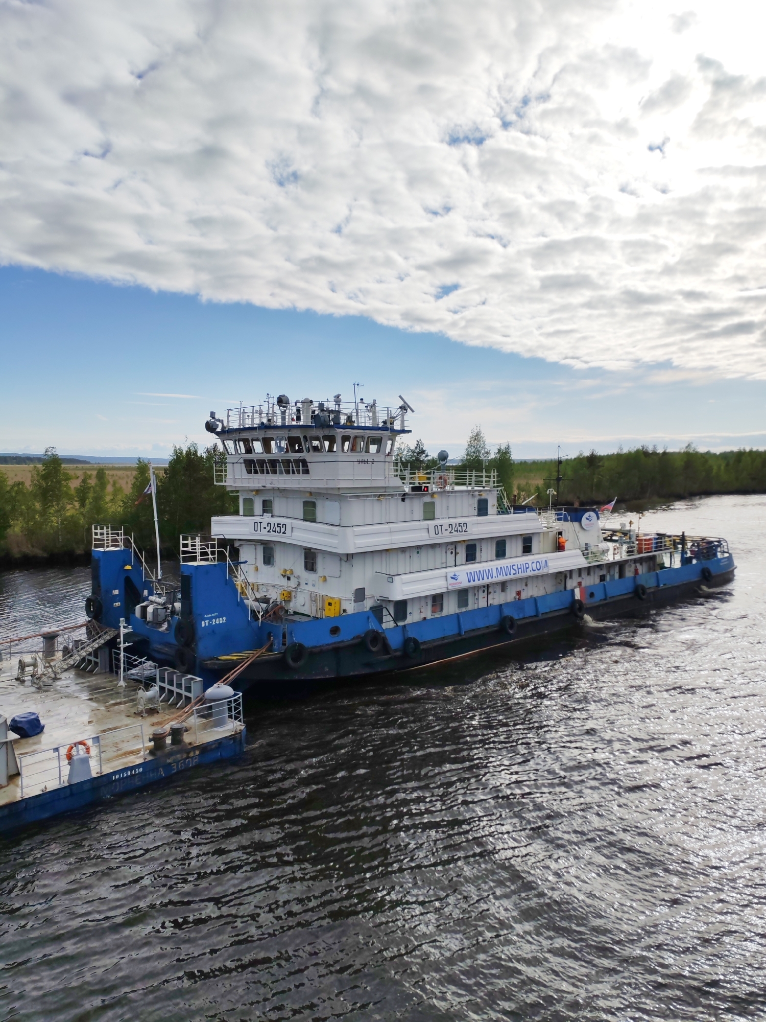 A blue and white tugboat named "OT-2452," with the website "WWW.MWSHIP.COM" written on its side, navigates a dark, rippled river. Attached to the tugboat is a smaller barge. The river is flanked by green vegetation and trees. Above, a sky of white clouds and blue is visible. The image was taken at the city of Ozernoe Ust'e, Russia.