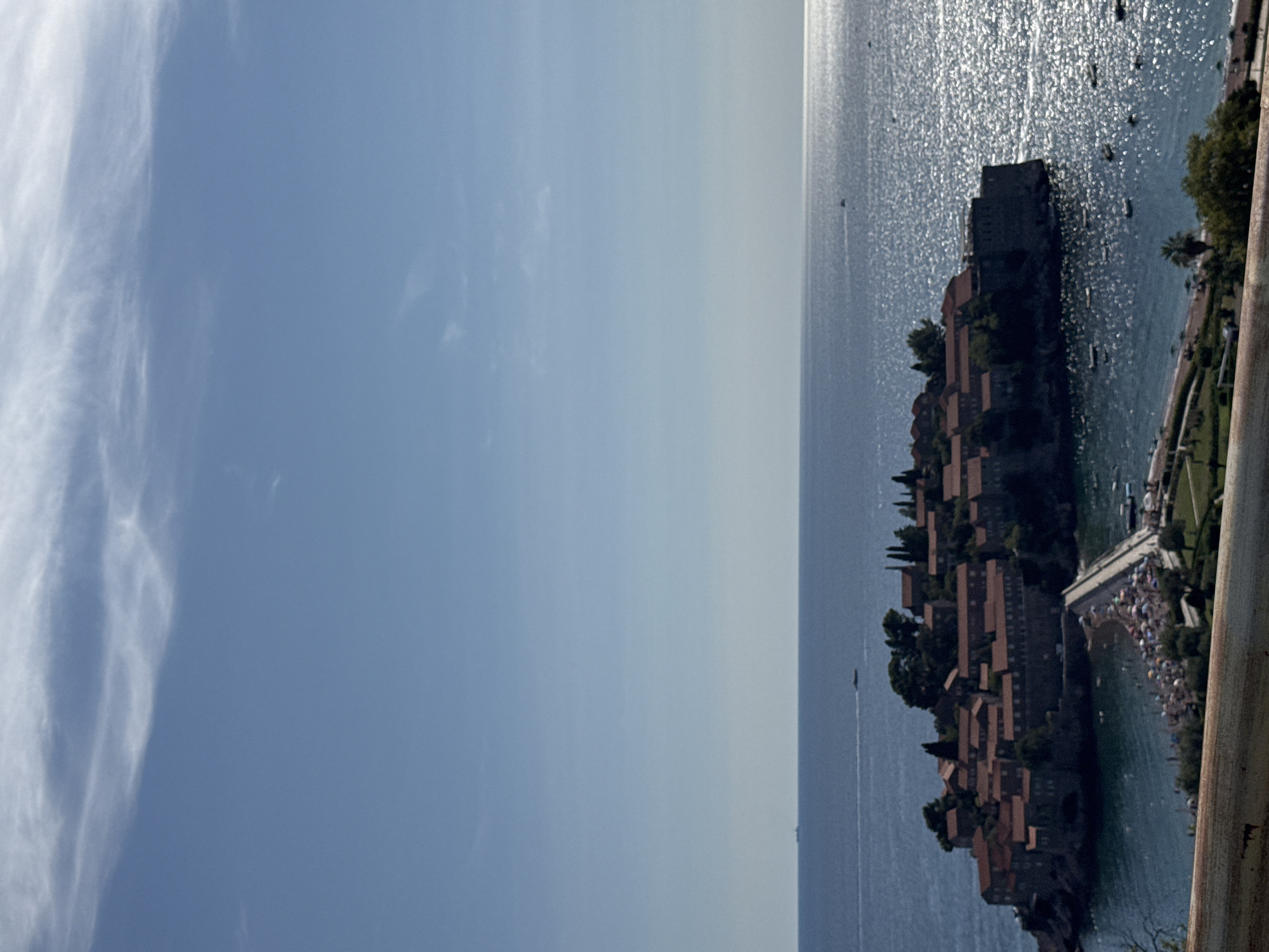 The image depicts a panoramic view of Sveti Stefan, a coastal town and islet in Montenegro, bathed in bright daylight under a clear blue sky with scattered white clouds. The focal point is the historic islet of Sveti Stefan, characterized by its cluster of terracotta-roofed buildings, appearing as a densely packed village atop a rocky outcrop. A narrow causeway, also lined with numerous beach umbrellas and a congregation of people, connects the islet to the mainland. The shimmering Adriatic Sea surrounds the islet, reflecting the sunlight intensely. Several small boats are visible scattered across the water. The foreground features a section of the coastline with lush greenery and a palm tree. The overall scene conveys a serene and picturesque coastal landscape.