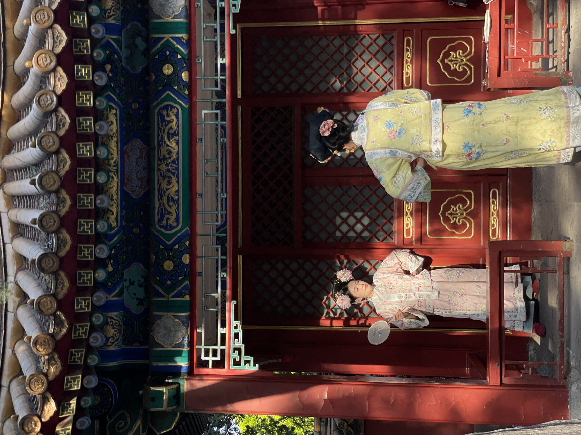 Two individuals in traditional Chinese garments are positioned within or against a building's doorway. The building is characterized by red and ornate architectural details, including lattice-patterned doors. A woman in a yellow floral dress is standing, with her back partially facing the camera, while another woman in a pink floral dress is seated. The building's architectural elements consist of intricate blue, green, and gold details on the left side of the frame, along with rows of cylindrical components at the top left. The scene is illuminated by sunlight, with a glimpse of greenery visible at the bottom of the image. The location is the Dongcheng District, China.