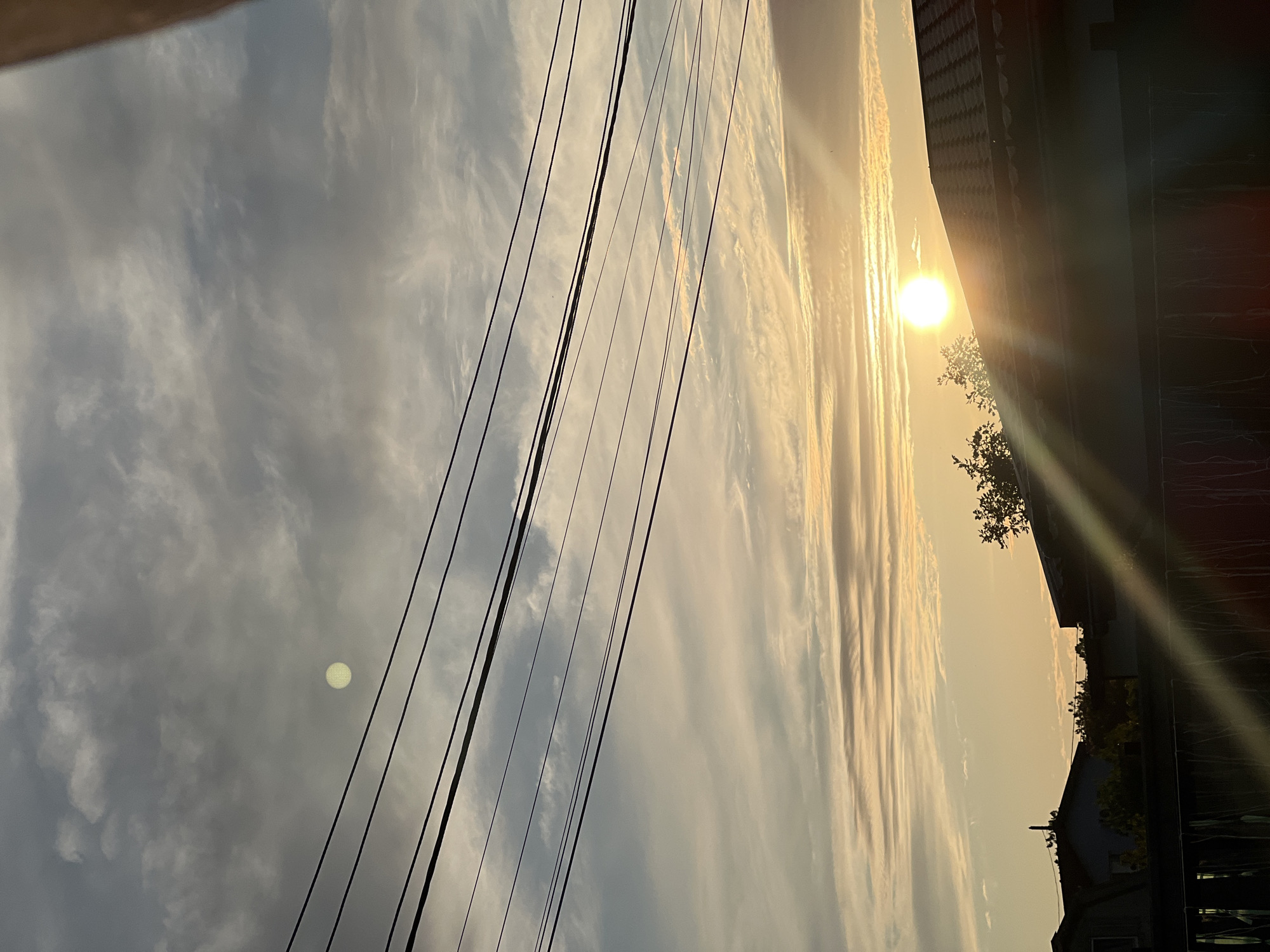 A low-angle, outdoor, vertical shot shows a sky filled with clouds during sunrise or sunset. Power lines stretch horizontally across the frame, and the sun is visible, casting a bright glare. In the lower portion of the image, the silhouettes of rooftops and trees are visible against the sky. The scene is set in Unknown, Unknown.