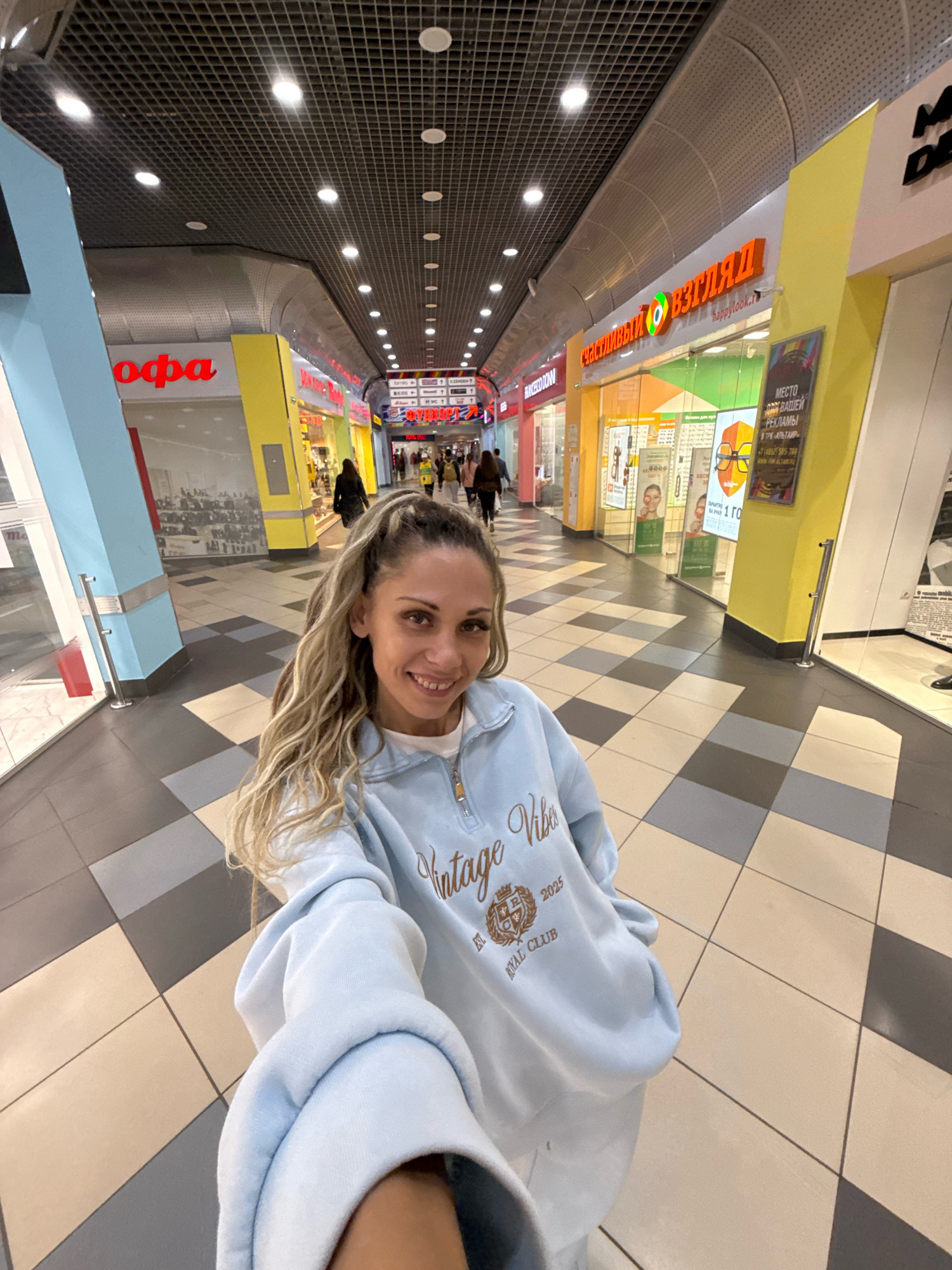 The image is a selfie taken by a young woman with long, blonde, wavy hair and a friendly smile, standing in a brightly lit shopping mall corridor. She is wearing a light blue sweatshirt with a "Vintage Vibes Royal Club 2025" logo. The corridor has a checkered floor with alternating light grey and beige tiles. On either side of the corridor are storefronts with colorful signs. To the left, a storefront has a large blue and red sign with "офа" in red Cyrillic letters. Further down the corridor, there are shops with yellow and green signage. The ceiling features a grid-like structure with recessed lighting. Several other shoppers are visible in the background, walking down the corridor. The scene appears to be inside a commercial establishment in Sosnovtsy, Russia, indicated by the Cyrillic text on the signage.