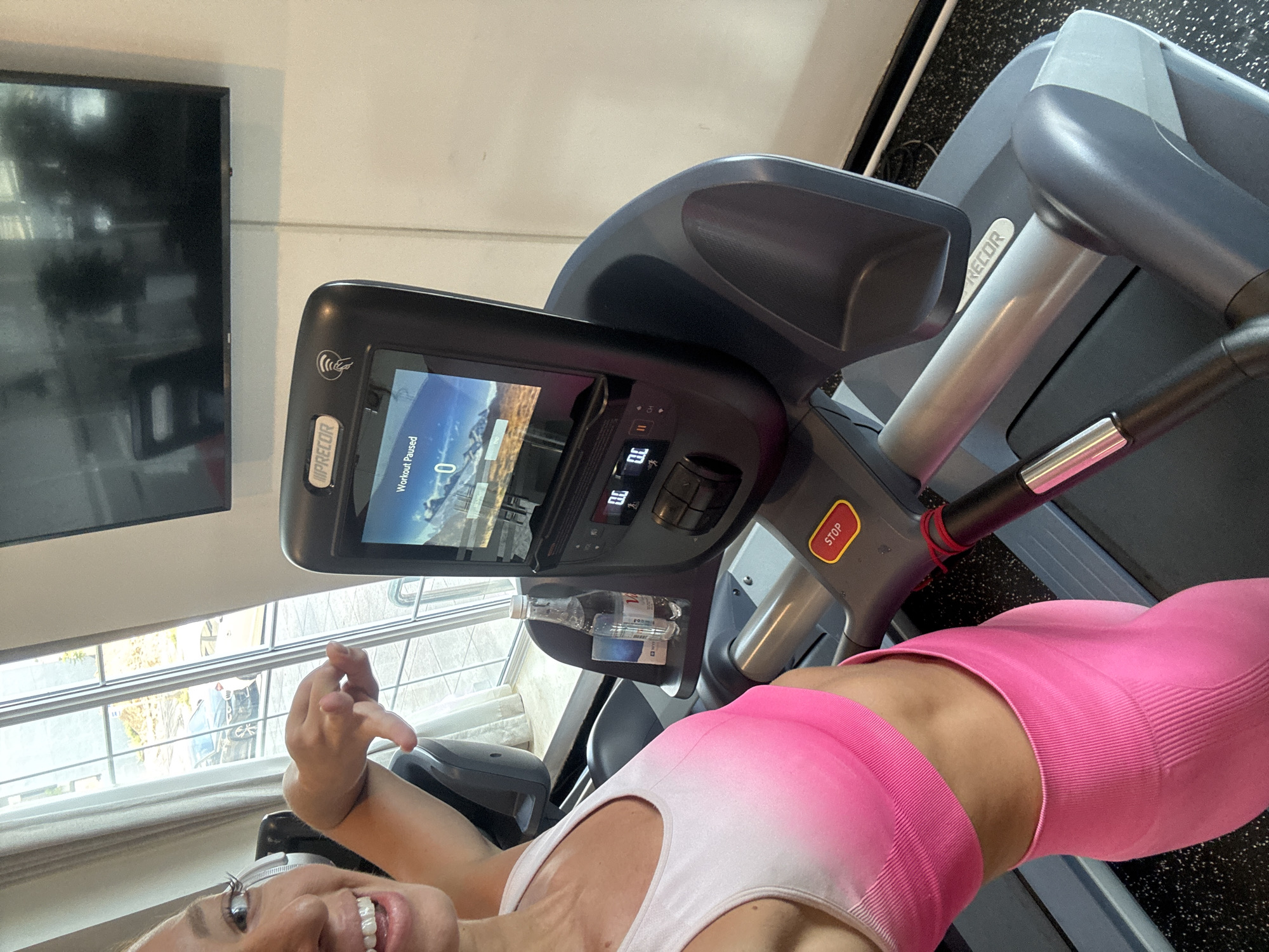 A woman in pink athletic wear is in the foreground, positioned on a Precor treadmill. The treadmill's console displays "Workout Paused" with a value of "0." A water bottle is visible in a holder on the treadmill. A television is mounted on the wall in the background, to the left of the treadmill. A window with visible blinds is located behind the woman, letting in daylight. The environment appears to be a gym or fitness center with rubberized flooring. There is no contextual information to suggest that the location is in Đà Nẵng, Vietnam.