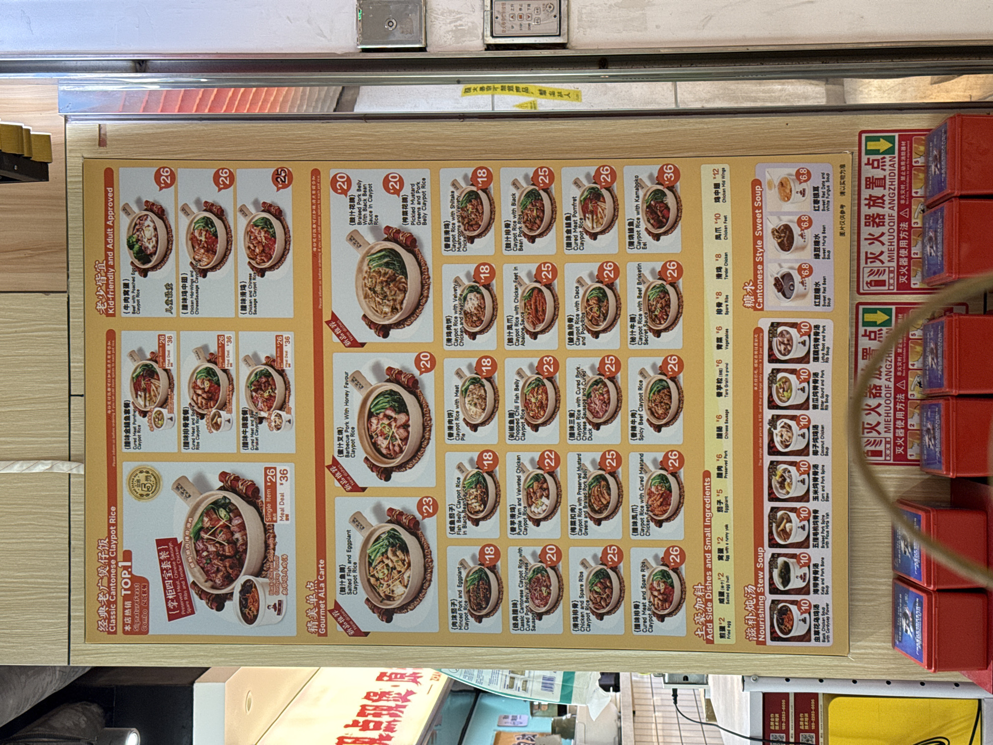 The image shows a menu board for a restaurant specializing in Cantonese claypot rice in Liwan District, China. The board displays colorful photos of various claypot rice dishes, their ingredients (such as meats, vegetables, and seafood), and their prices. Sections include "Classic Cantonese Claypot Rice," "Gourmet ALa Carte," "Nourishing Stew Soup," and "Cantonese Style Sweet Soup." Pricing information is included, listing both single-item and meal-deal options. The board also notes that the restaurant is "Kid-friendly and Adult Approved." Contact information for technical training and brand partnerships is provided at the bottom. A fire extinguisher and instructions for its use are visible on the right edge.