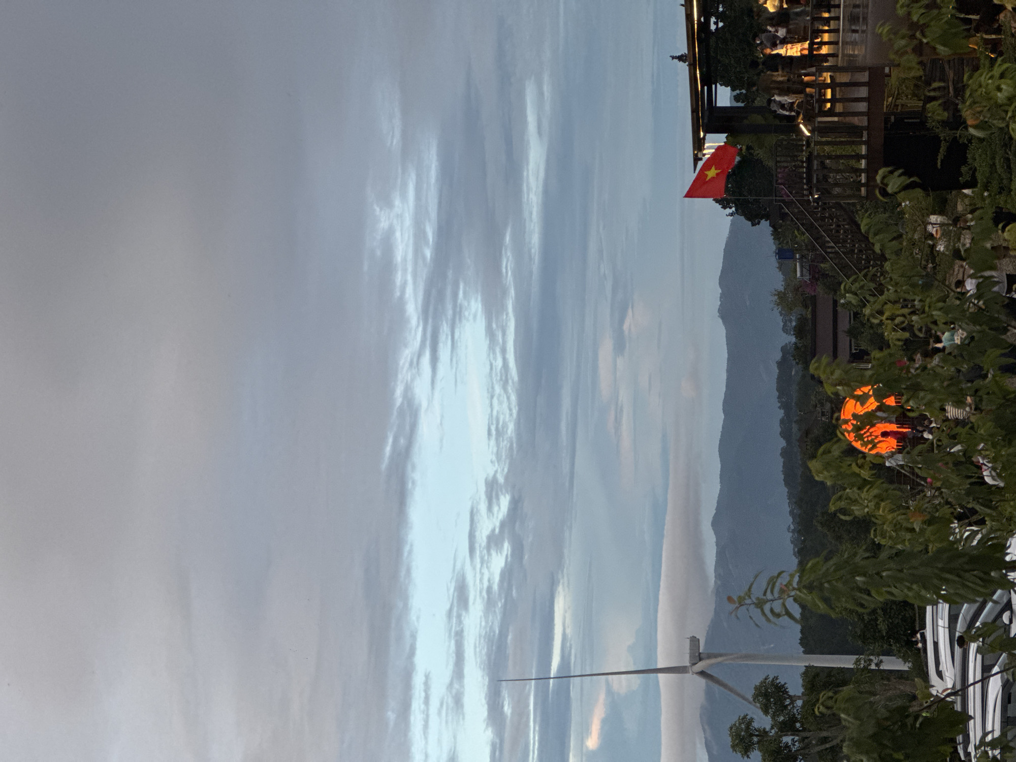 Captured in Phát Chi, Vietnam, the image features a landscape dominated by a partly cloudy sky and mountainous terrain in the background. A Vietnamese flag is prominently displayed on a wooden structure reminiscent of a restaurant or viewing platform. The foreground includes lush greenery, and an orange circular light fixture is visible amidst the vegetation. Additionally, a wind turbine is noticeable near the lower part of the frame.