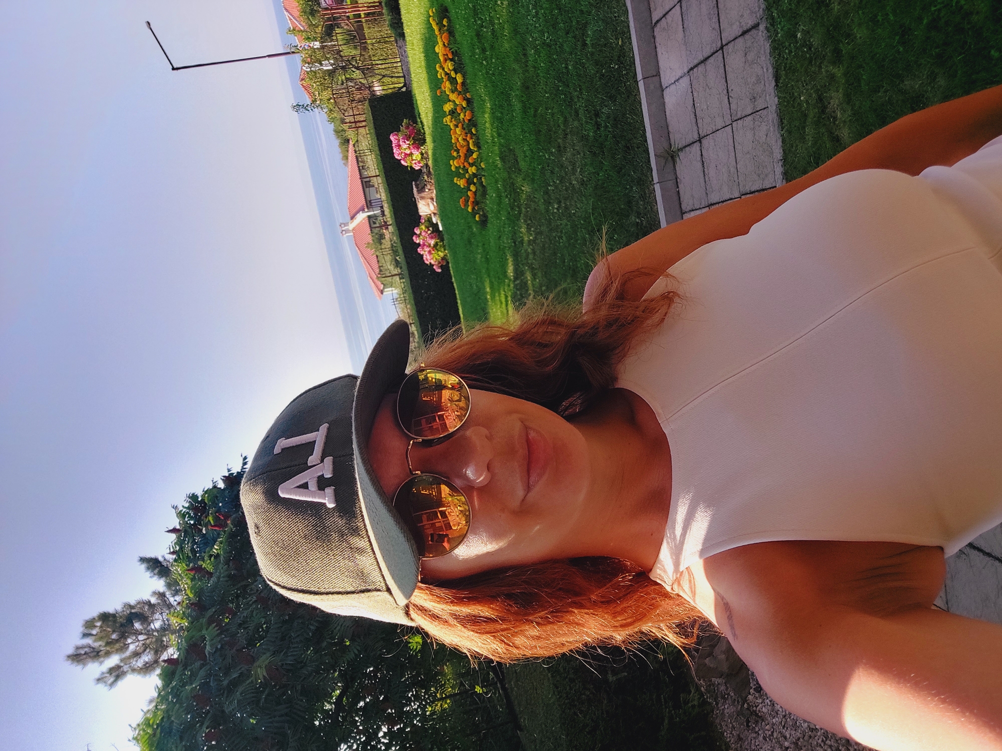 The image shows a woman with red hair wearing a dark green baseball cap with white letters "AJ" and round sunglasses. She is wearing a white sleeveless top. She is standing outdoors in front of a green lawn with flower beds containing yellow and orange flowers. In the background, there are houses with red roofs and the sea. The scene appears to be in a residential area in Sozopol, Bulgaria. The lighting suggests it is daytime, possibly late afternoon due to the warm glow.