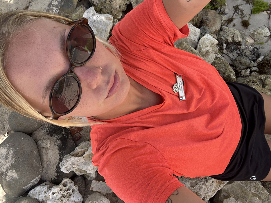 The image presents a high-angle selfie of a blonde individual, identified as "LORIANA JUNIOR CLUB" by a name tag featuring a French flag emblem pinned to a coral-colored V-neck shirt. The individual wears dark oval sunglasses and a small gold nose ring in the left nostril, complemented by black athletic shorts and a visible partial tattoo on the left arm. Reflections in the sunglasses depict a body of water, sand, and an individual in a green garment. The immediate environment is a rocky shoreline composed of varying sizes of grey and white coral fragments, intermingled with patches of sand. The scene is situated in Sainte-Anne, France.