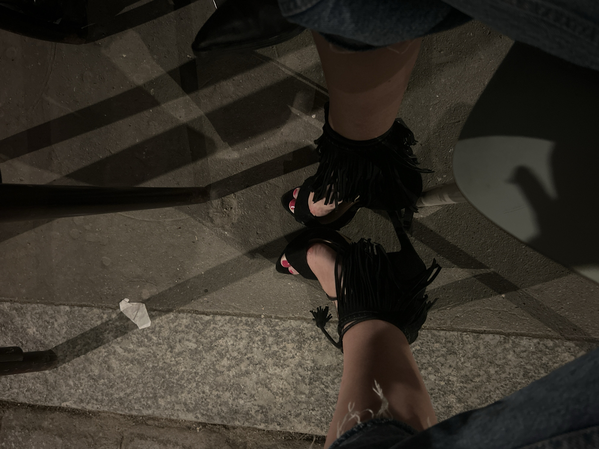 The image captures a scene on an urban street. The subject's legs and feet are prominent, with the person wearing open-toed, high-heeled sandals featuring fringed embellishments. The toes are painted with red nail polish. The subject is wearing what appears to be denim jeans. The location seems to be outdoors, on a paved surface that appears to be a city street or sidewalk. There are shadows cast across the pavement, suggesting the presence of overhead structures. In the background, there is a white piece of trash on the ground. The setting is likely an urban area.