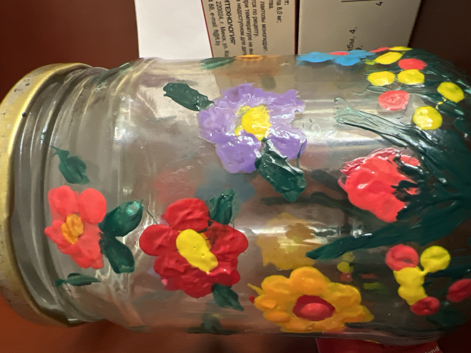 The image shows a glass jar with painted flowers. The jar has a gold lid. The flowers are in various colors including red, yellow, and purple, and are painted in a somewhat abstract, child-like style. Some green leaves are also painted around the flowers. In the background, a white box with Russian text is visible. The text on the box appears to be pharmaceutical information, including an address in Minsk, Belarus: "220024, г. Минск, ул. Ко" which translates to "220024, Minsk, K. street". This indicates that the jar is likely located in an urban environment, possibly a home or a shop, in Minsk, Belarus, given the label on the box. The lighting suggests it is indoors.