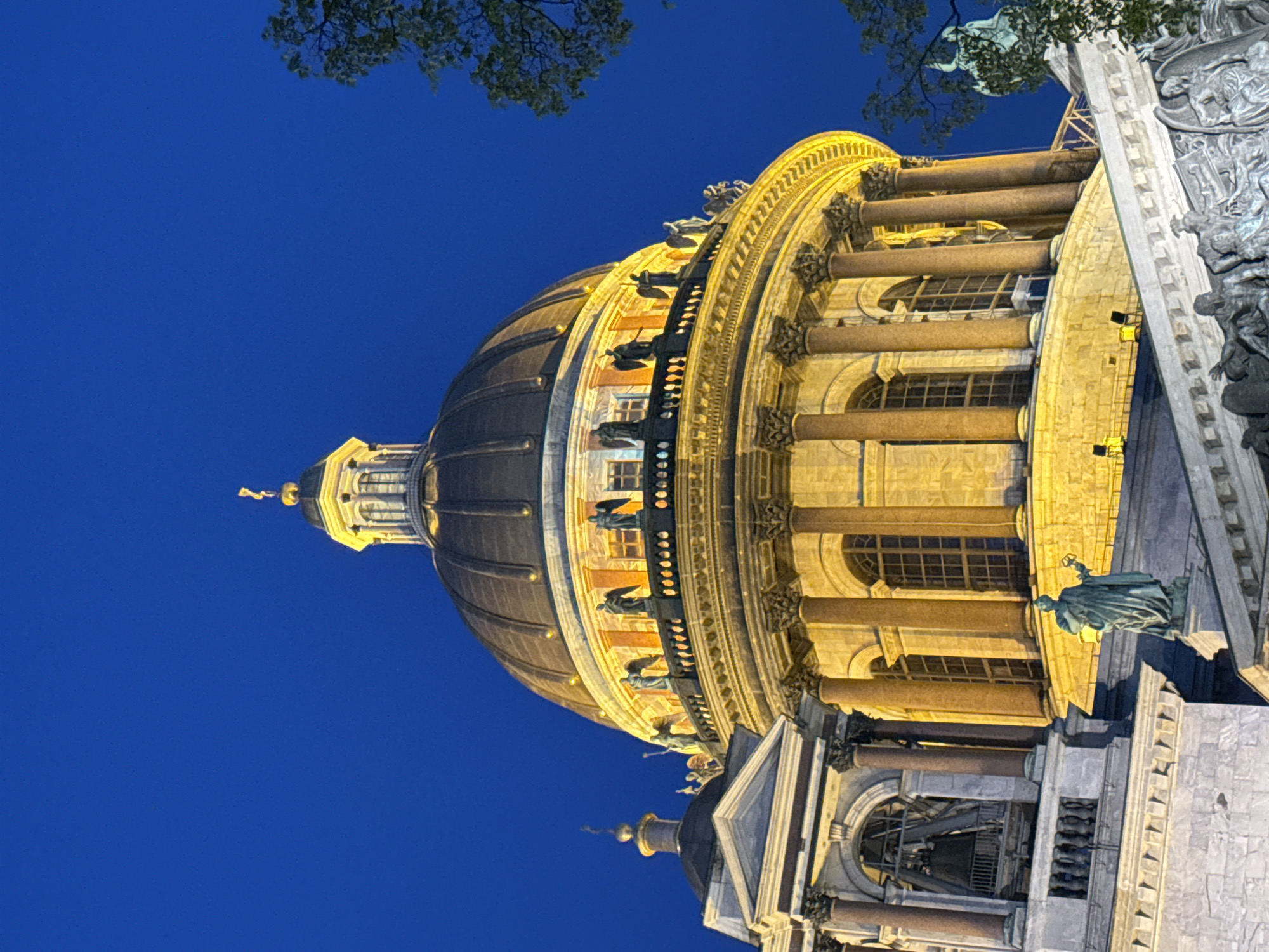 The image, which is rotated 90 degrees clockwise, presents a striking night view of Saint Isaac's Cathedral in Saint Petersburg, Russia. The grand architectural masterpiece is bathed in warm, golden artificial light, contrasting sharply with the clear, deep indigo blue sky, indicating late twilight or nighttime.
The central feature is the cathedral's massive, bronze-colored dome, crowned by a smaller golden cupola and a delicate spire at its apex. Encircling the base of the main dome is a brightly illuminated drum, adorned with several large arched windows. A balustrade runs along the top of this drum, populated by numerous statues of winged figures, likely angels, which stand out prominently due to the strong illumination.
Below the dome's drum, a magnificent colonnade of polished reddish-brown granite columns wraps around the structure. These towering columns, likely Corinthian in style, support a robust entablature and frame sections of the building with additional arched and rectangular windows. The artificial lighting dramatically emphasizes the intricate architectural details, the carvings, and the textures of the stone and metalwork.
On the lower right side of the image, beneath the colonnade, a large, dark statue of a robed figure, possibly a saint, stands prominently on a pedestal. To its right, a richly detailed sculptural relief panel, depicting multiple figures in high relief, adorns the building's facade. Green foliage from a tree extends into the upper left corner of the frame, and another branch is visible towards the upper right, adding a natural element to the urban architectural scene. No specific activity is depicted, and no visible text is present. The overall impression is one of grandeur, majesty, and historical significance, beautifully showcased under the evening sky.
