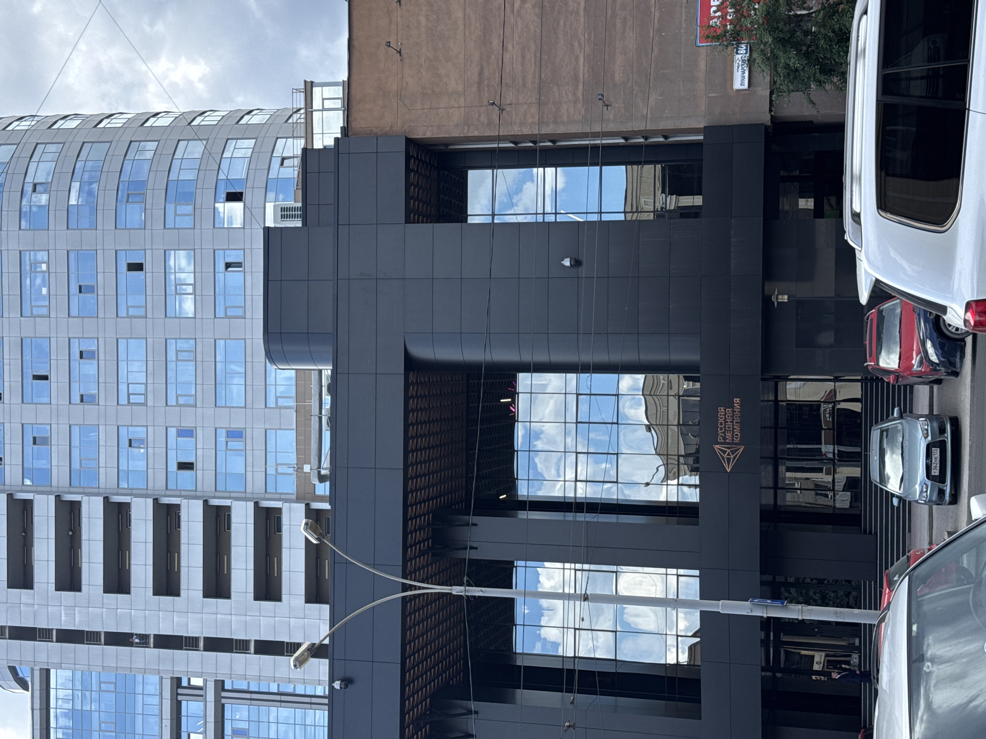 The image shows a modern office building in Yekaterinburg, Russia, with reflective glass facades. A prominent sign on the dark-colored lower section of the building reads "РУССКАЯ МЕДНАЯ КОМПАНИЯ" (Russian Copper Company). The building is situated on a street with parked cars, including a silver sedan with a license plate visible, and a white SUV. The sky is overcast with clouds. The architecture suggests a contemporary urban setting.