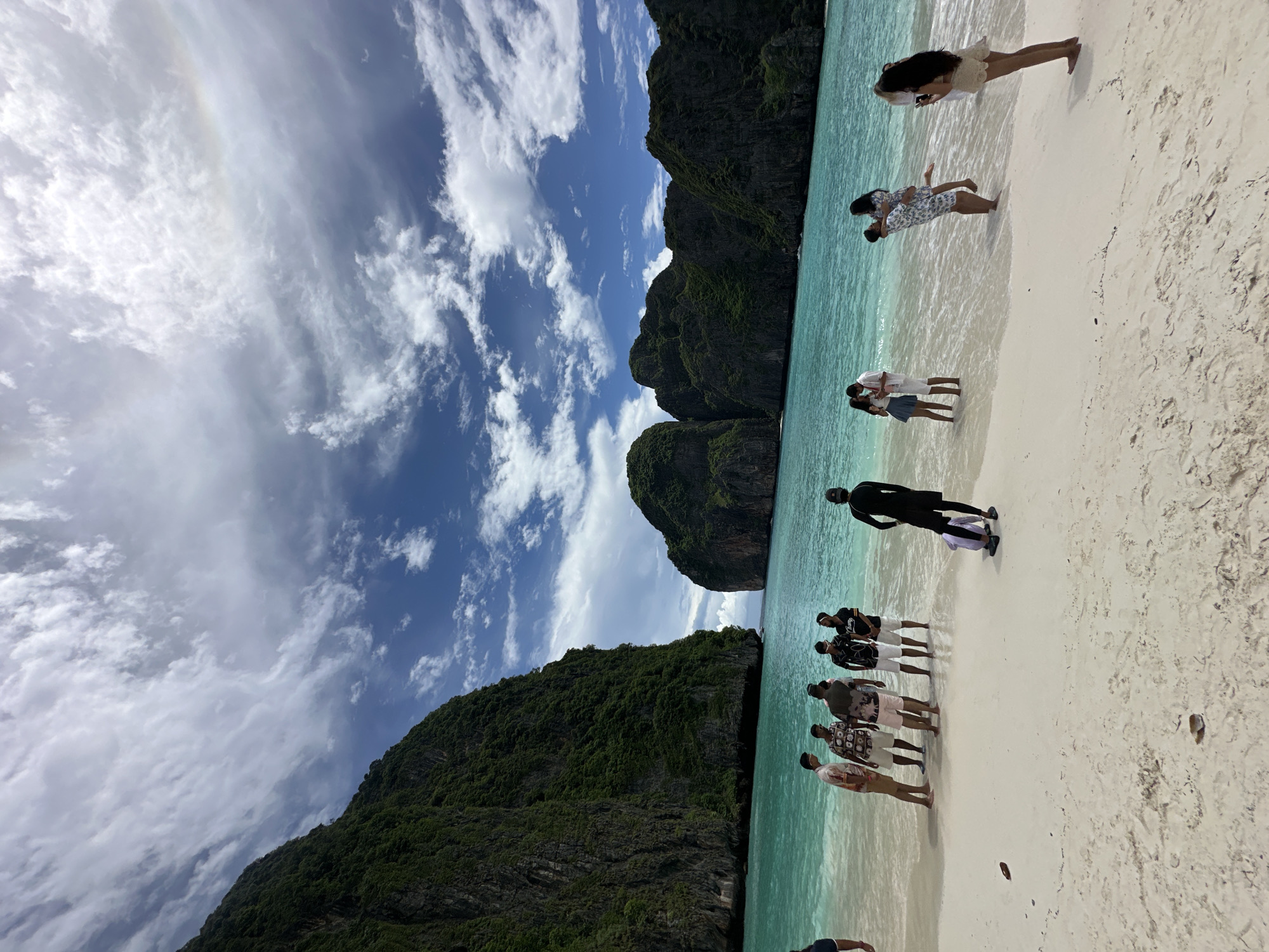 The image depicts a wide view of Maya Bay in Krabi, Thailand, a renowned tourist destination. The bay is characterized by its stunning turquoise waters and towering limestone cliffs covered in lush greenery. In the foreground, a white sandy beach is occupied by several groups of people.
On the left side of the beach, a group of men, possibly tourists, are gathered, some standing and some talking. Further to the right, a person dressed in black is seen carrying a rolled-up mat or bag. In the middle ground, several couples are interacting in the shallow water and on the sand. One couple is engaged in a romantic pose, with a man lifting a woman. Another couple is standing close together, and a woman is taking a photograph with her phone.
The sky above is a vibrant blue, dotted with white, fluffy clouds, and a faint rainbow is visible. The overall scene conveys a sense of leisure and natural beauty, typical of a tropical beach resort.
