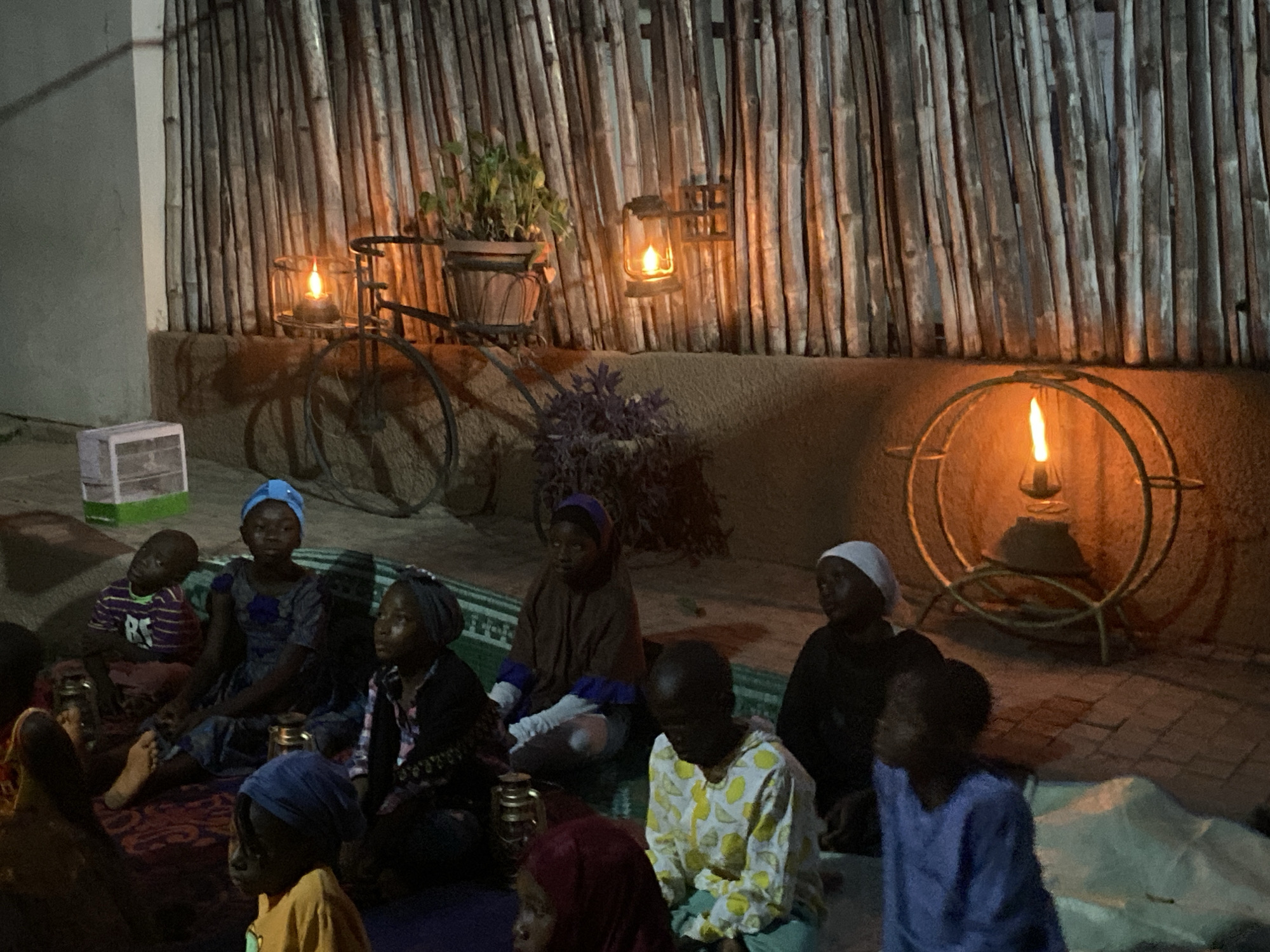 In Barden Yana, Nigeria, a group of children are sitting on mats outdoors at night. The lighting is low, with several lanterns providing illumination. In the background, there is a wall constructed of vertical wooden poles, decorated with plants and lanterns. The children are wearing various clothing items, including head coverings. They appear to be gathered together, possibly listening to a story or participating in an activity. There are small metal lanterns next to the people on the mats.