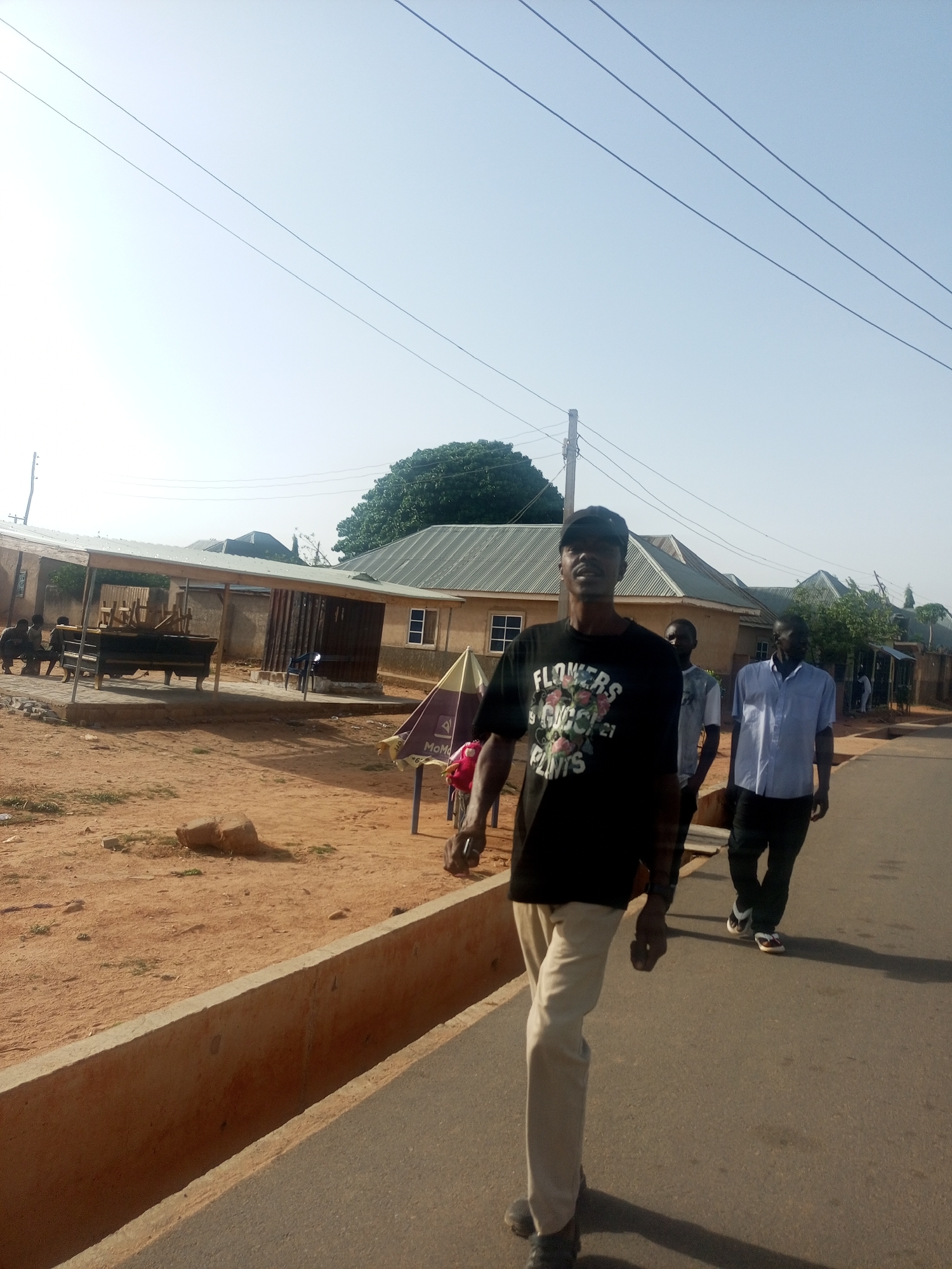 The image shows a street in an unknown city. A man wearing a black t-shirt with "FLOWERS Gucci 21 PLANTS" printed on it, light brown pants, and a black cap is walking toward the camera. Two other men are walking behind him. In the background, there are buildings with corrugated metal roofs, a partially visible parasol, and a dirt ground on the left side of the road. Overhead power lines are visible against a light blue sky.
