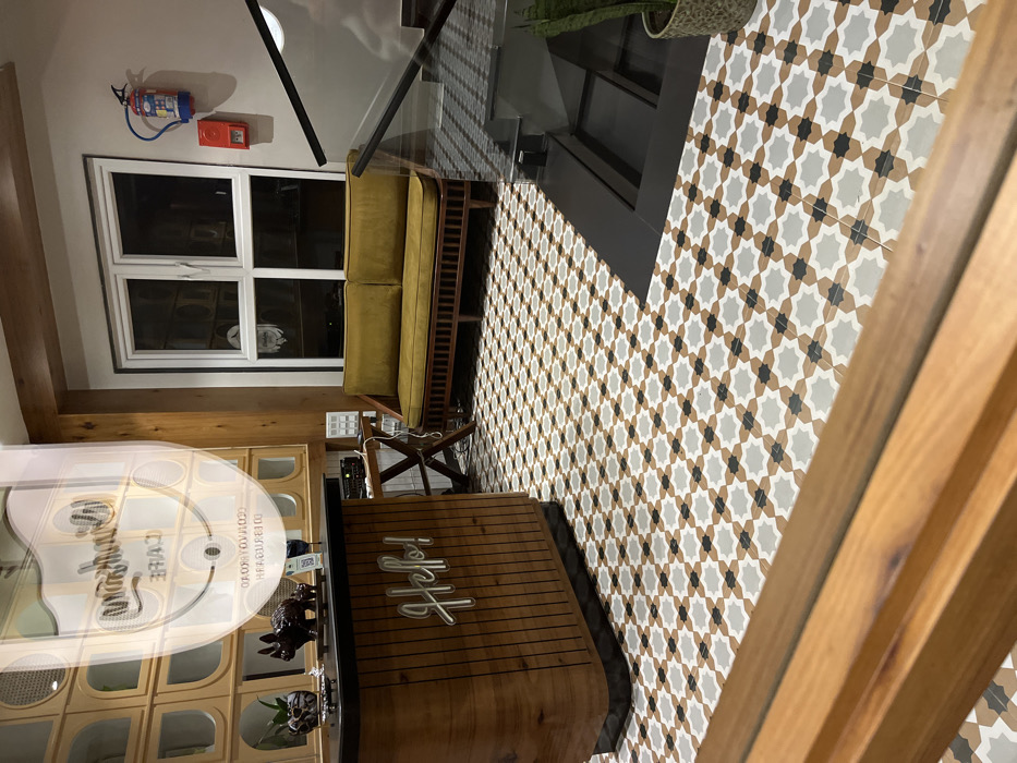 This image captures the interior of a commercial establishment, likely a cafe or lounge, featuring modern decor and geometric patterns. The perspective is tilted, looking down into the space.
A dominant feature is the floor, which is covered in intricate geometric tiles. These tiles form a repetitive star-like pattern using light grey, beige, and black colors. A dark, possibly elevated, section of flooring divides the patterned area.
To the left of the frame, a white-framed window is visible on a light-colored wall. Above the window, a red fire extinguisher and a small red box (likely a fire alarm) are mounted. Below the window, a contemporary yellow sofa with cushions and ribbed wooden details offers seating. Next to the sofa, a small wooden table with an X-shaped base holds what appears to be electronic equipment.
In the mid-ground and foreground, a wooden reception or service counter is prominent. It features vertical wooden slats and a curved design. A neon sign spelling "Heft" is affixed to the front of this counter. To its left, part of a wooden display unit with multiple cubbies or shelves can be seen. A clear glass barrier or railing extends across the mid-ground, leading to a wooden railing or banister on the far right.
Reflections on the glass surfaces within the image provide additional information, showing the name "Sungrow Cafe" and the address "COMMERCIAL ROAD, DIBRUGARH." This detail, combined with the user-provided context of "Dibrugarh, India," confirms the location. The scene appears unoccupied, suggesting it might be captured during off-hours.