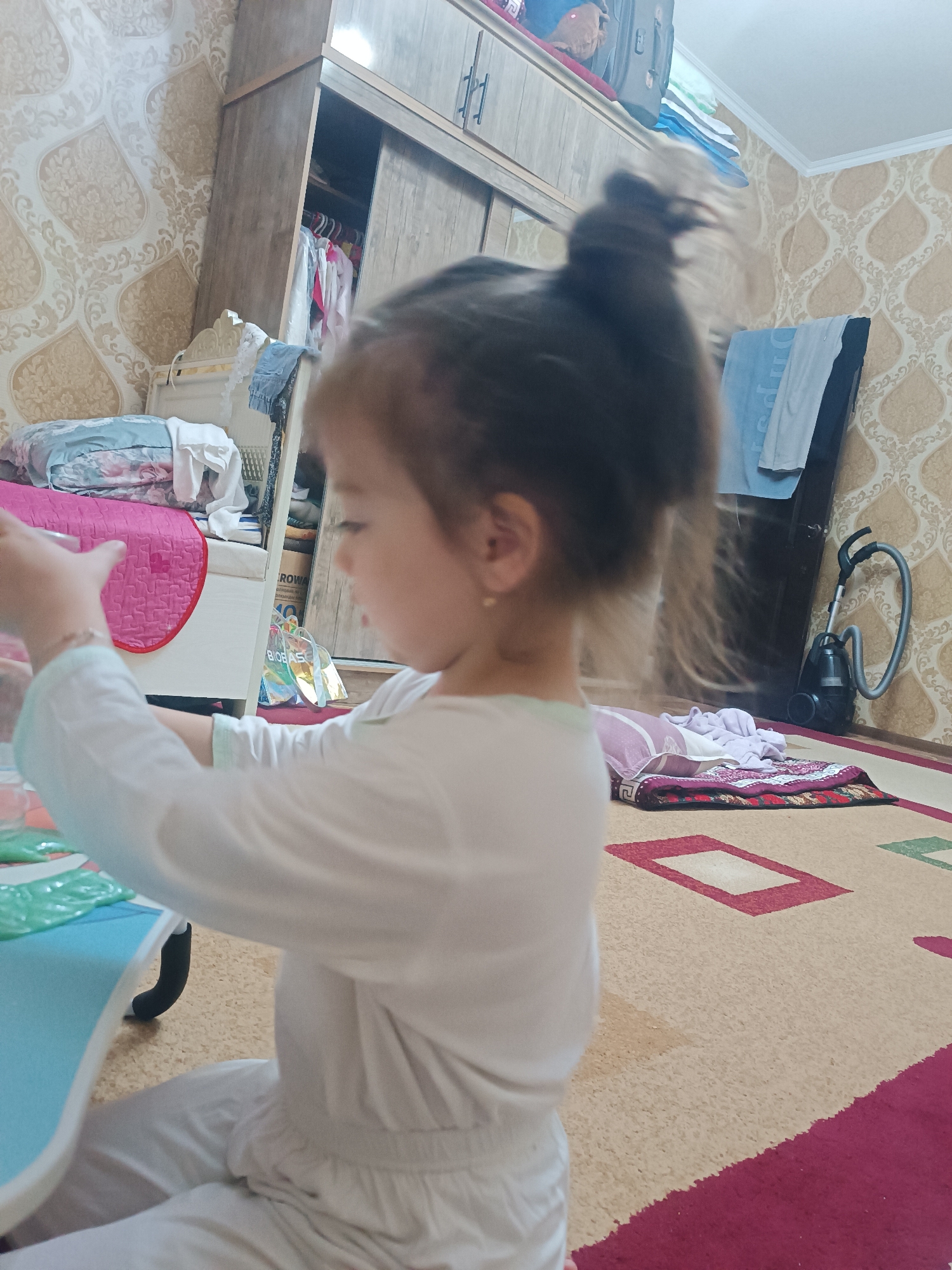 A young girl with her hair in a bun, wearing a white outfit, is seated at a blue table, her hands near a collection of green plastic packets. She is positioned in a room with patterned wallpaper and a large wooden wardrobe. A vacuum cleaner is visible in the background, near a doorway. A rug with geometric patterns covers the floor. The scene appears to be an indoor residential setting in Navoiy City, Uzbekistan.