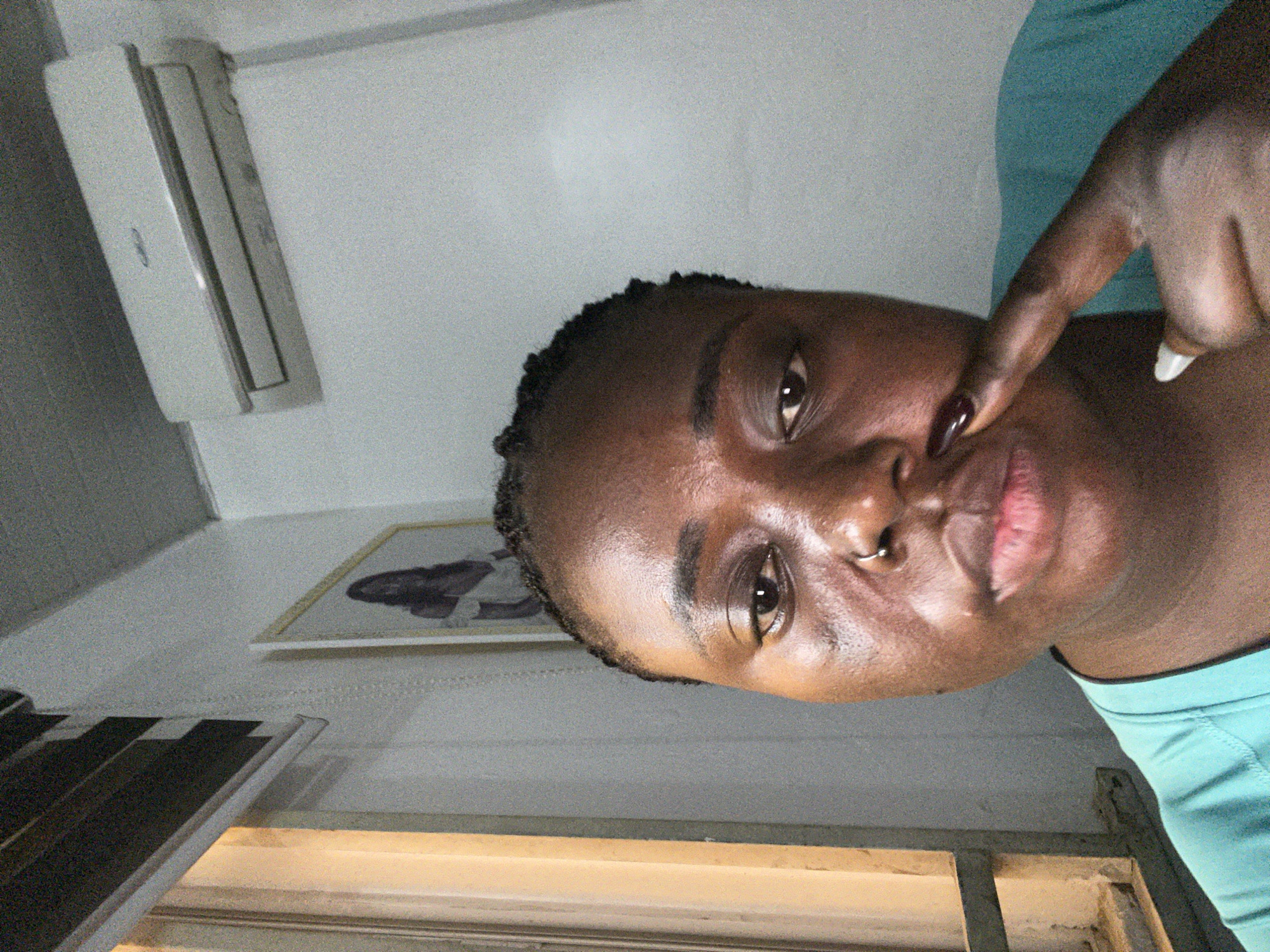 In Abuja, Nigeria, a person with dark skin and braided hair is taking a selfie. They are wearing a turquoise t-shirt and have a nose ring. Their right index finger, with dark nail polish, is touching the tip of their nose. In the background, a framed picture of a woman hangs on the wall, and an air conditioning unit is mounted above it. A window with blinds is visible to the left. The lighting suggests an indoor setting, possibly a room.