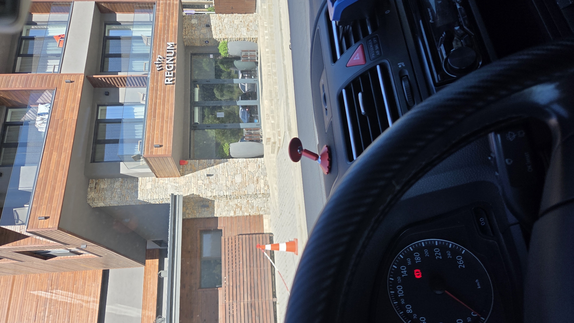 The image shows the exterior of a modern building with a sign that reads "REGNUM". The building features a combination of wood and stone facade, with balconies on multiple levels. In the foreground, the interior of a car is visible, with a steering wheel and dashboard. A red traffic cone is placed on the sidewalk in front of the building, and a white rope barrier is visible. There are also two large potted plants outside the building's entrance. The scene appears to be a street view in an urban or semi-urban setting.