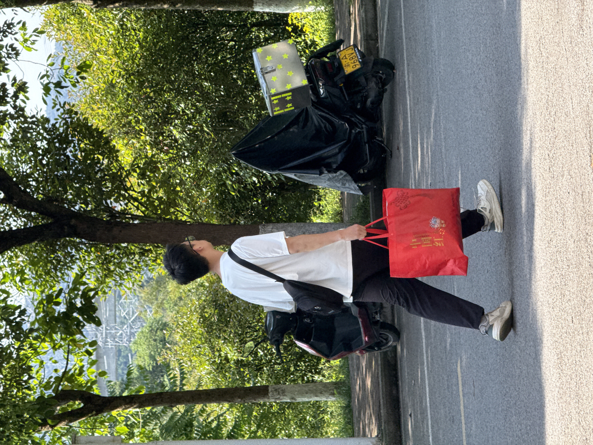The image captures a sunny daytime scene in Jiangjin, China, featuring a person walking on a paved road alongside a tree-lined area. The individual, viewed from behind and slightly to the right, is a male wearing a white t-shirt, black pants, white sneakers, and glasses. A black shoulder bag is slung across their body, and they are carrying a bright red shopping bag in their right hand.
To the left of the person, a dark-colored scooter or motorcycle is parked. It has a yellow license plate, clearly displaying "渝 A 968VJ," where "渝" is the abbreviation for Chongqing, which encompasses Jiangjin. A silver-toned delivery box, decorated with numerous yellow stars and horizontal yellow stripes, is mounted on the back of the scooter, which is partially covered by a black tarp.
The ground consists of a gray asphalt road on the left, with a lighter, textured pavement on the right, possibly a sidewalk or shoulder. Shadows are cast across the ground, indicating bright sunlight. In the background, beyond the dense green trees and foliage, a large metal structure, possibly a power line tower or part of a bridge, is visible against the bright sky, suggesting an urban or semi-urban landscape. The red shopping bag prominently features Chinese characters, including "中国" (China) and "茅台" (Maotai), implying its contents are related to Maotai liquor.