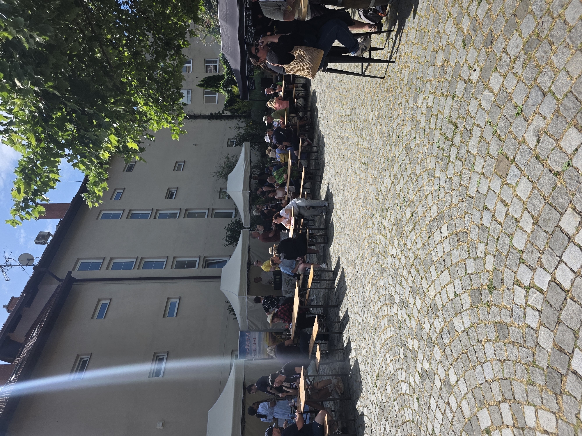 The image depicts an outdoor scene at an event in an unknown city. Several people are seated at long wooden tables, some are under white tents. A building with multiple windows is in the background, partially obscured by a tree. The ground is paved with cobblestones. Some individuals are visible at the tables, appearing to be engaged in conversation and consuming food or beverages.