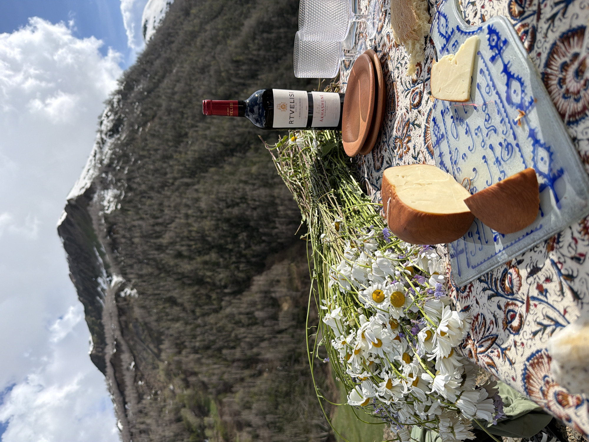 This is a medium shot, eye-level, outdoor, daylight, still life photograph of a picnic spread on a table with a mountainous landscape in the background. The table is covered with a colorful, patterned tablecloth. In the foreground, on the left, is a large bouquet of white daisies with yellow centers, some small purple flowers, and green stems. To the right of the flowers, there are two pieces of cheese on a blue and white patterned cutting board. One piece is a wedge, and the other is a thin slice. A large half-wheel of cheese, with a darker rind, is positioned to the left of the cutting board. Behind the cheese and flowers, there is a bottle of red wine labeled "RTVELISI" and "AKHASRENI," standing upright. To the right of the wine bottle are two clear, textured glasses. In the background, a steep, tree-covered mountain slope rises, with patches of snow visible on the upper reaches. The sky above is blue with scattered white clouds. The location is identified as Ushguli, Georgia. There are no people or animals present.