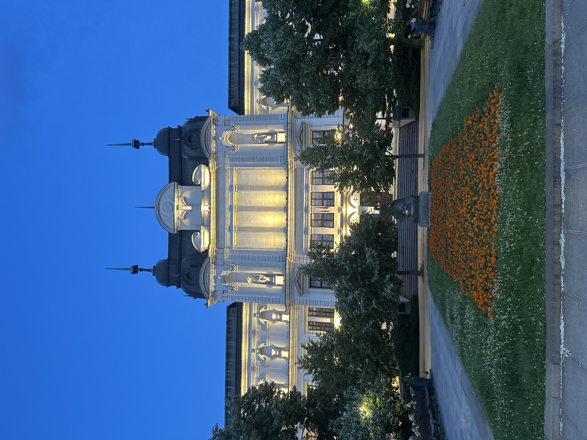 A white building with ornate architecture is lit from within and by external lights, highlighting its classical features. The building has a grand central facade with statues in niches and a dark, domed roof topped with spires. In front of the building is a manicured lawn with a large flowerbed of orange flowers and a smaller section of white flowers. A statue of a seated figure is positioned in front of the flowerbed. Mature trees with full green foliage flank the building and the lawn. The scene is set against a clear, deep blue twilight sky. Benches are visible on either side of the lawn.