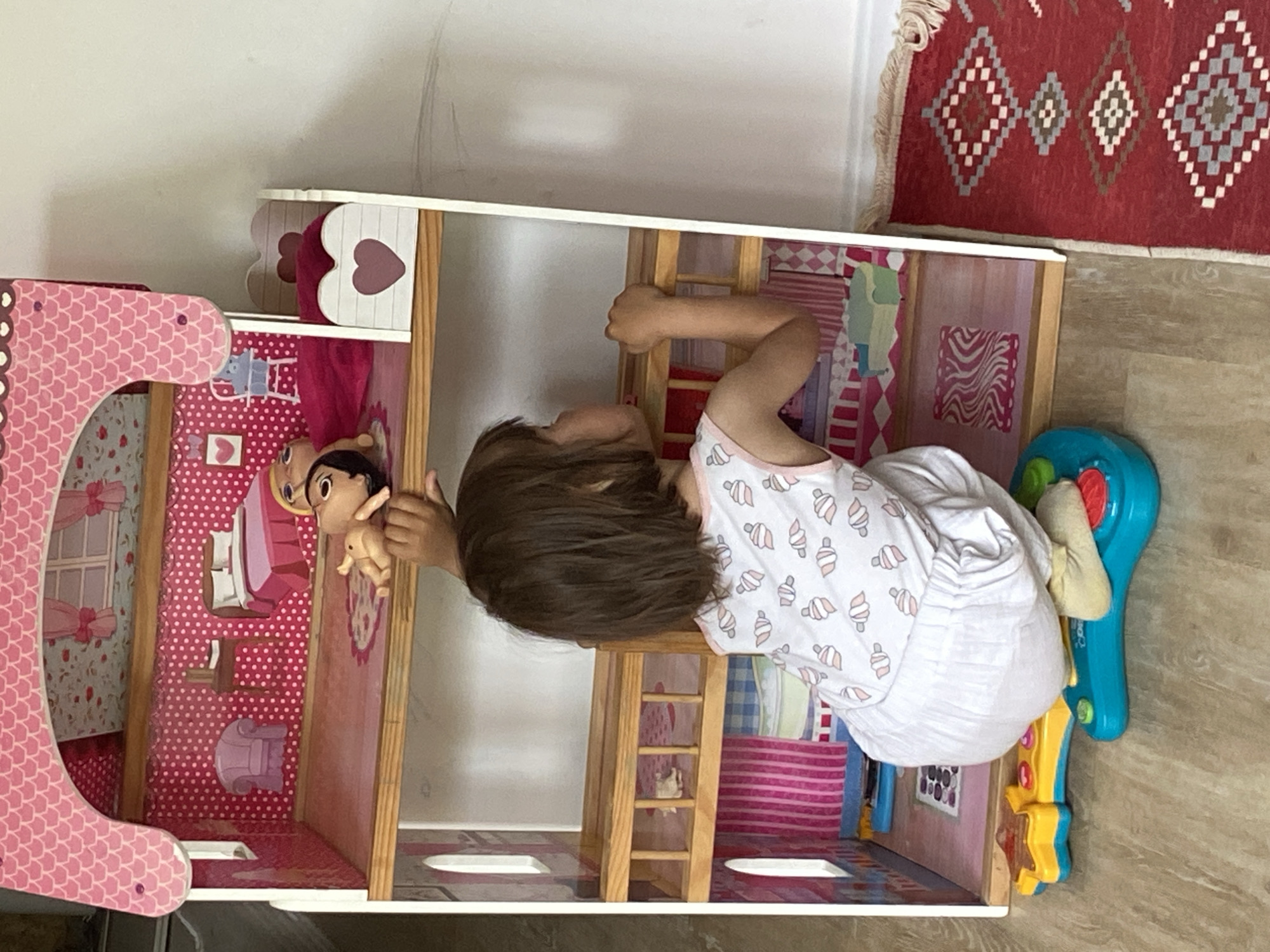 A child lies prone next to a dollhouse, holding a doll. The dollhouse is multi-story, pink and white, and filled with miniature furniture. A blue toy is positioned at the end of the dollhouse. The child wears a white shirt with ice cream cone designs. The floor is made of light-colored wood-look panels. A portion of a red rug with geometric patterns is visible near the top right of the frame. The location is likely indoors in Tyumen, Russia.