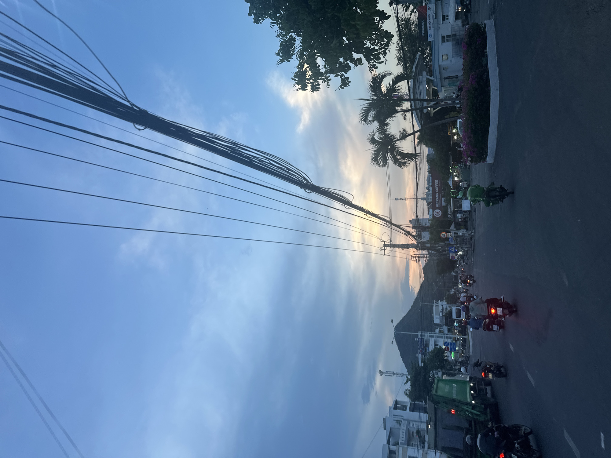 The image shows a street in Bac Nha Trang Ward, Vietnam, with power lines overhead. A mountain rises in the background. Motorbikes fill the street, some with visible headlights. A Metrang Coffee establishment is seen on the side of the road. Palm trees and other foliage line the sidewalk. Buildings are visible on both sides of the street. The sky is a mix of blue and cloud with light from the sunset. A green garbage truck is present.