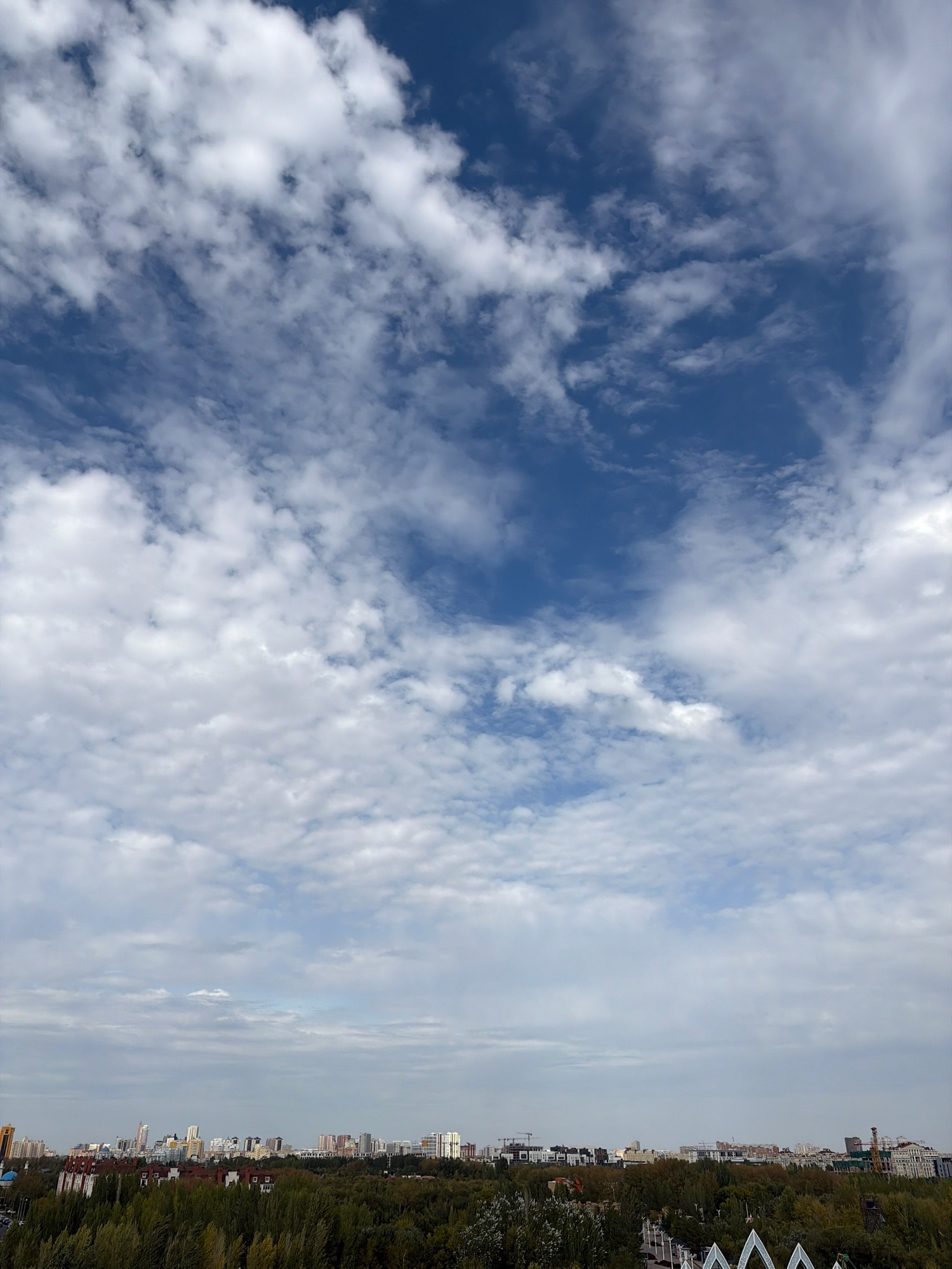 A panoramic view of a city skyline, likely Astana, Kazakhstan, is captured under a dynamic sky filled with a mix of white, fluffy clouds and darker, more wispy formations against a backdrop of deep blue. The cityscape stretches horizontally across the lower portion of the image, characterized by a dense array of buildings of varying heights and architectural styles. These include modern high-rises with glass facades, apartment complexes, and what appear to be construction cranes in the distance, suggesting ongoing urban development. A significant band of lush green trees forms a natural buffer between the city buildings and the viewer, indicating proximity to a park or natural area. The overall impression is one of a sprawling urban environment integrated with natural elements, under a vast, cloud-filled sky.